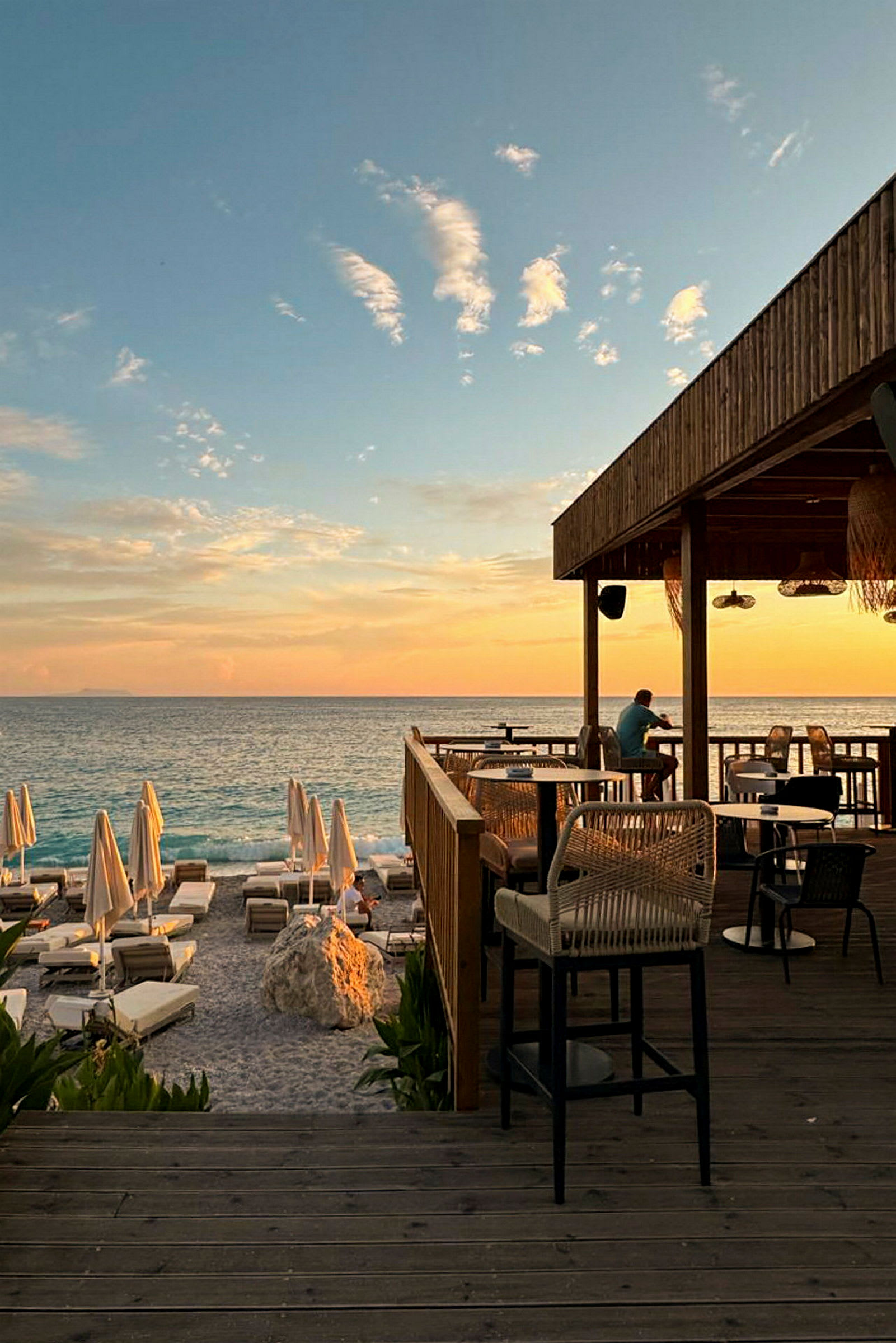 En solnedgång vid havet med en restaurangterrass och solstolar på stranden. (AI-genererad text)