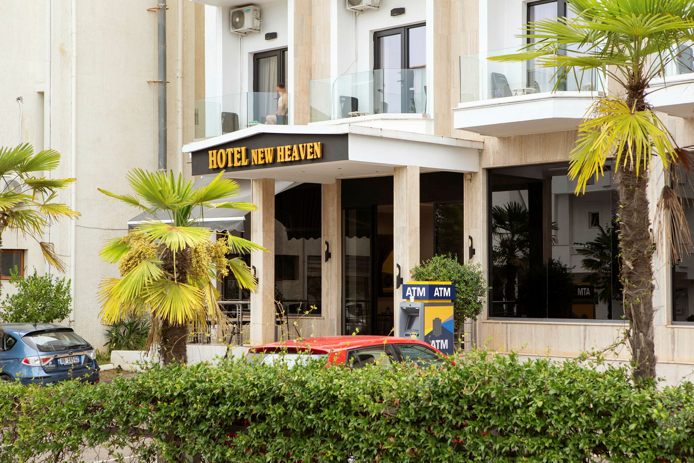 Byggnadens framsida med texten 'Hotel New Heaven', omgiven av palmer och en bankomat. (AI-genererad text)