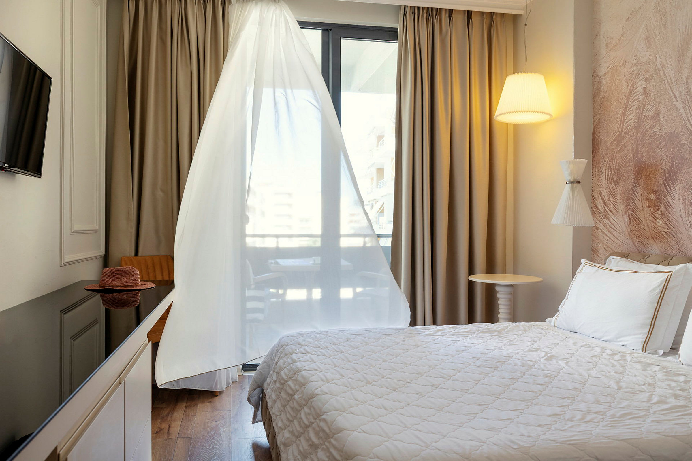 Ett hotellrum med en säng, TV, bord och stol, samt ett fönster med vita gardiner. (AI-genererad text)