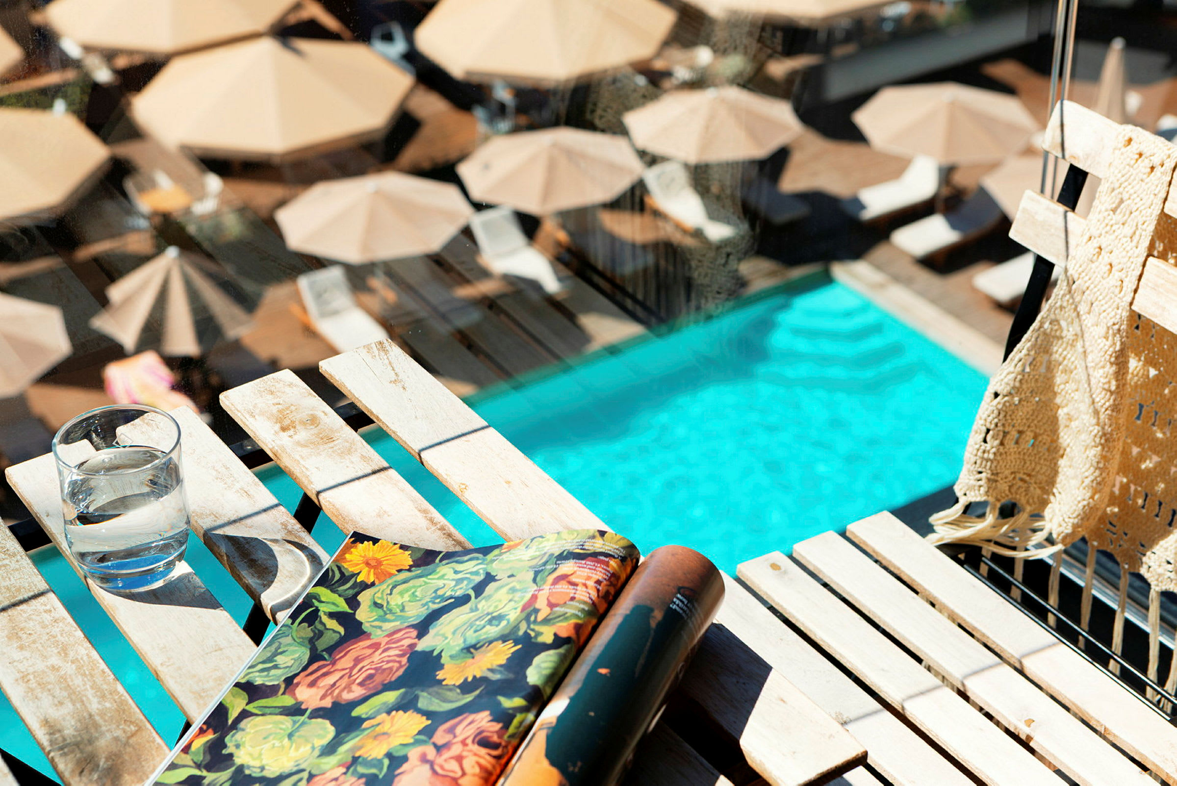 Bilden visar en pool omgiven av solstolar och parasoller, med fokus på ett bord med ett glas vatten, en bok och en filt. (AI-genererad text)