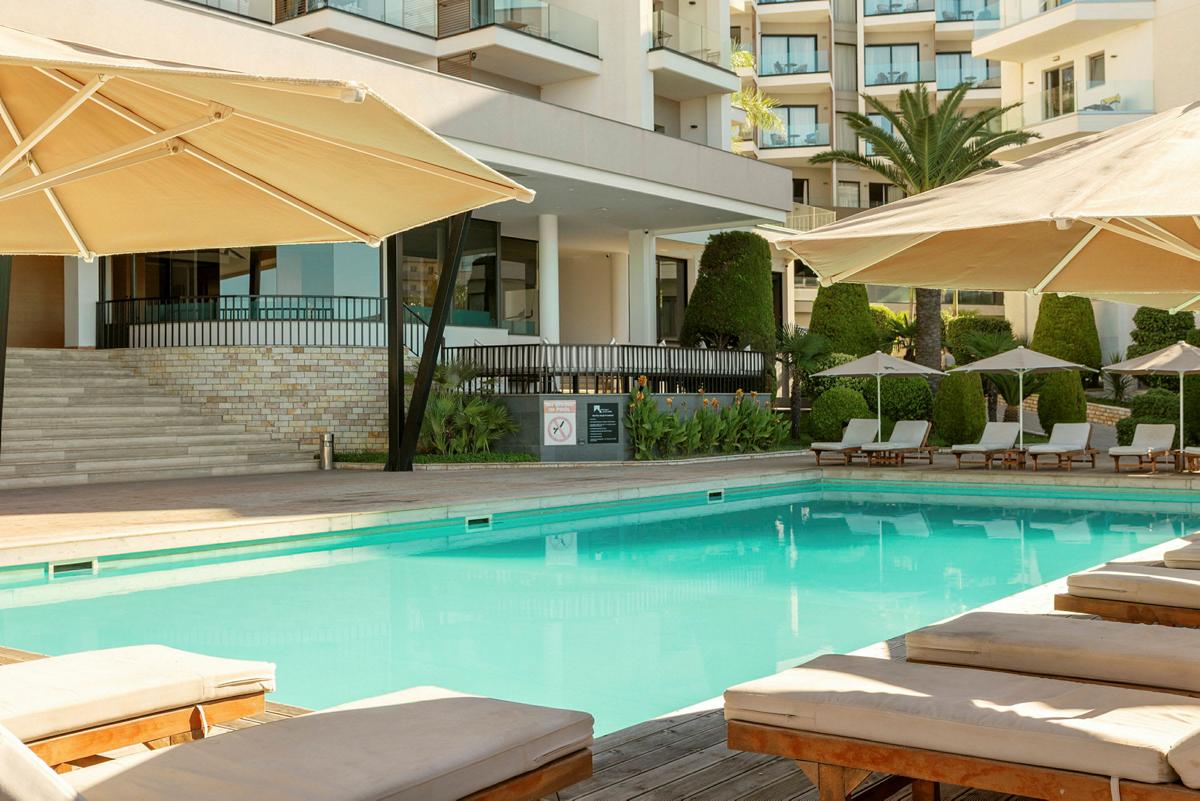 Bild av hotellpoolområde med parasoller och solstolar, byggnad i bakgrunden. (AI-genererad text)
