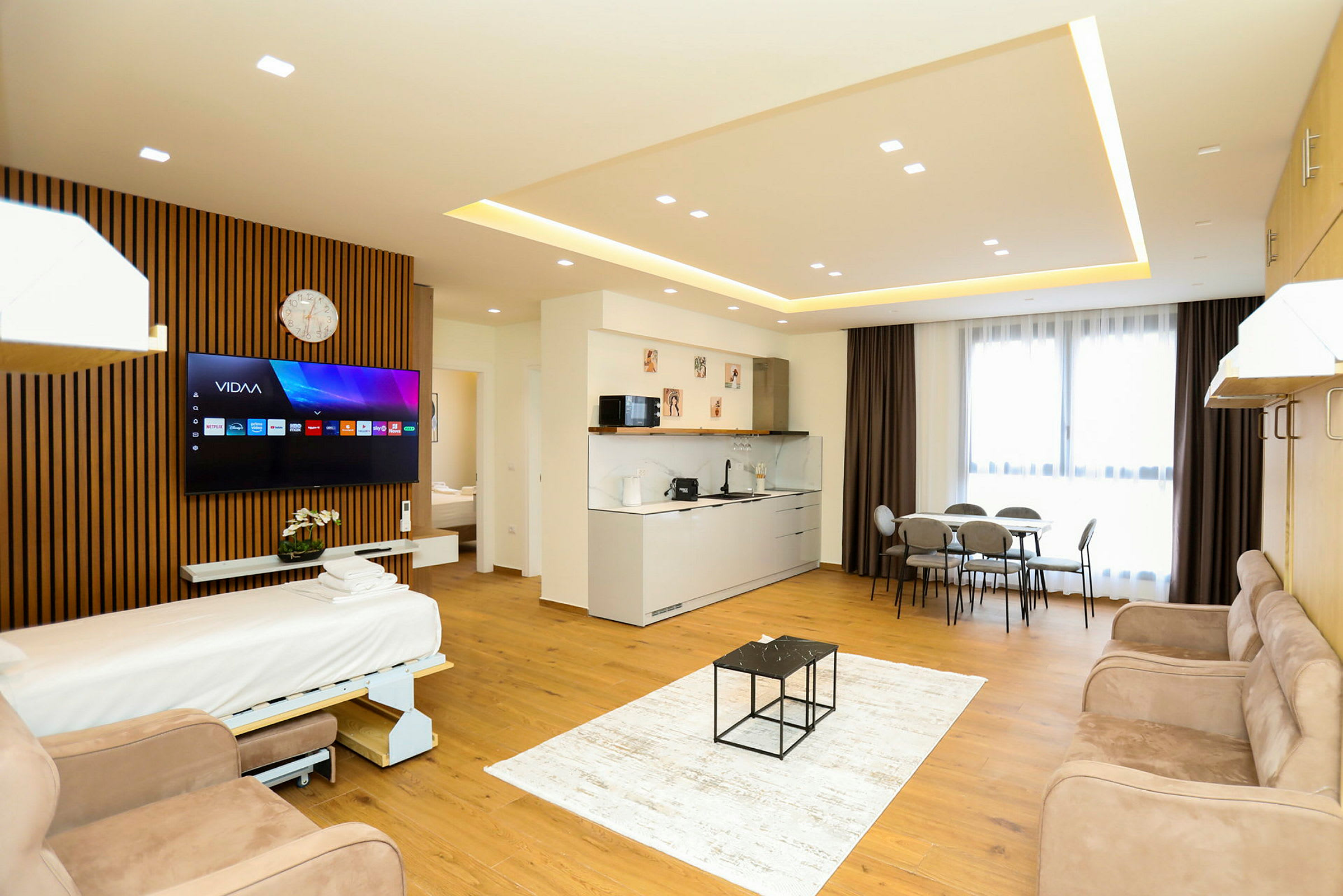 Ett modernt hotellrum med öppen planlösning inkluderande en bäddsoffa, TV, köksområde, matbord och stora fönster med gardiner. (AI-genererad text)