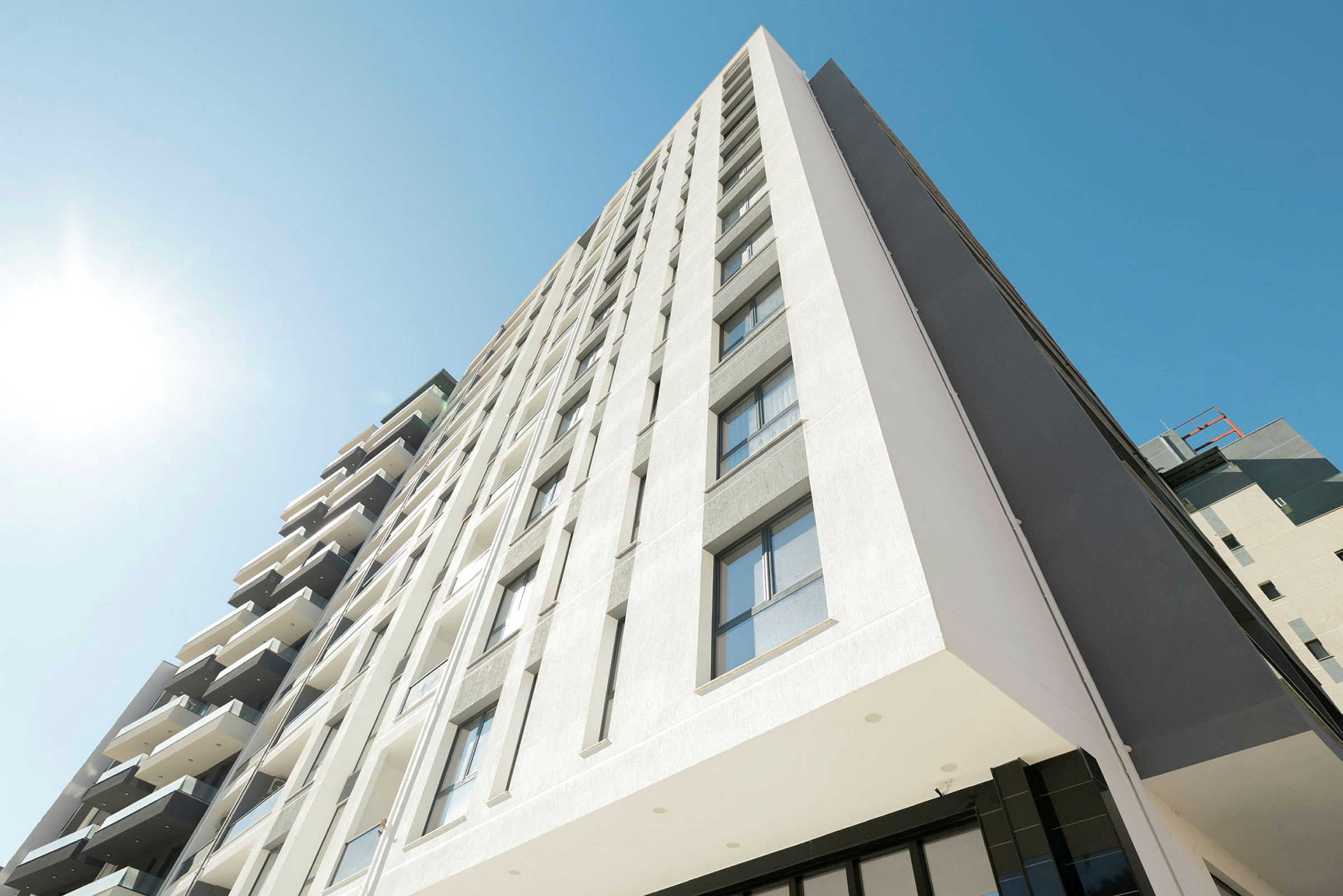 Bilden visar en modern byggnad med flera våningar, fasaden är ljus och har många fönster. En klar blå himmel och direkt solljus syns. (AI-genererad text)