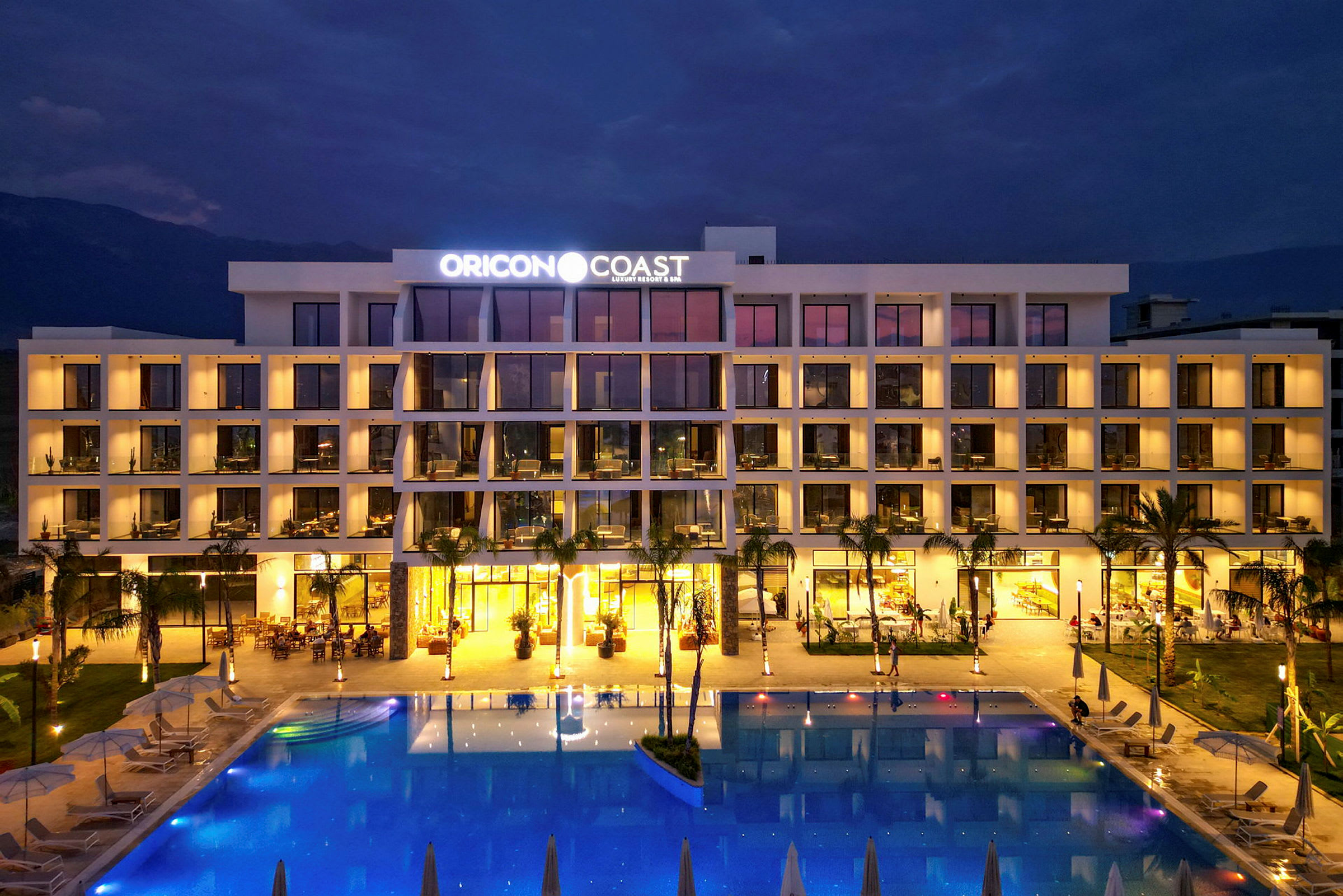Hotellfasad med pool framför, belyst på kvällen. (AI-genererad text)