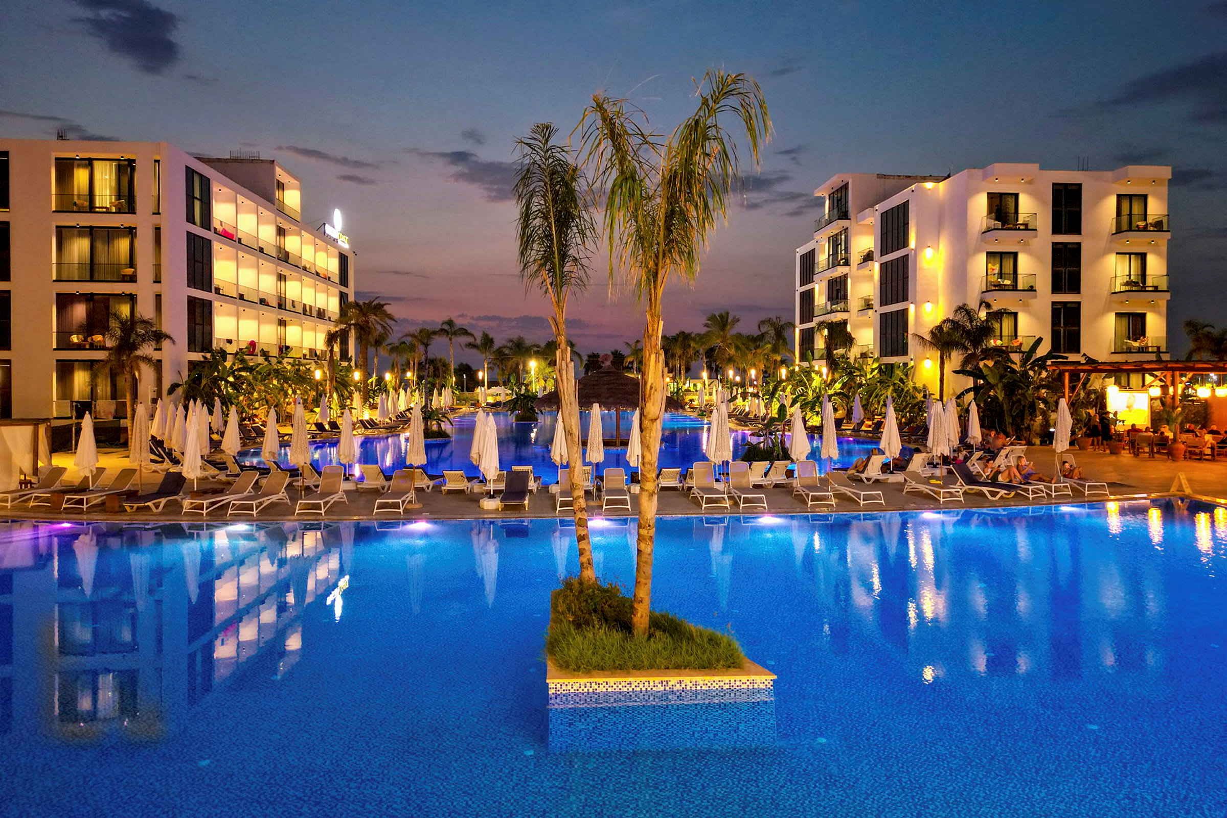 Kvällsvy över en upplyst hotellpool med parasoller och byggnader i bakgrunden. (AI-genererad text)