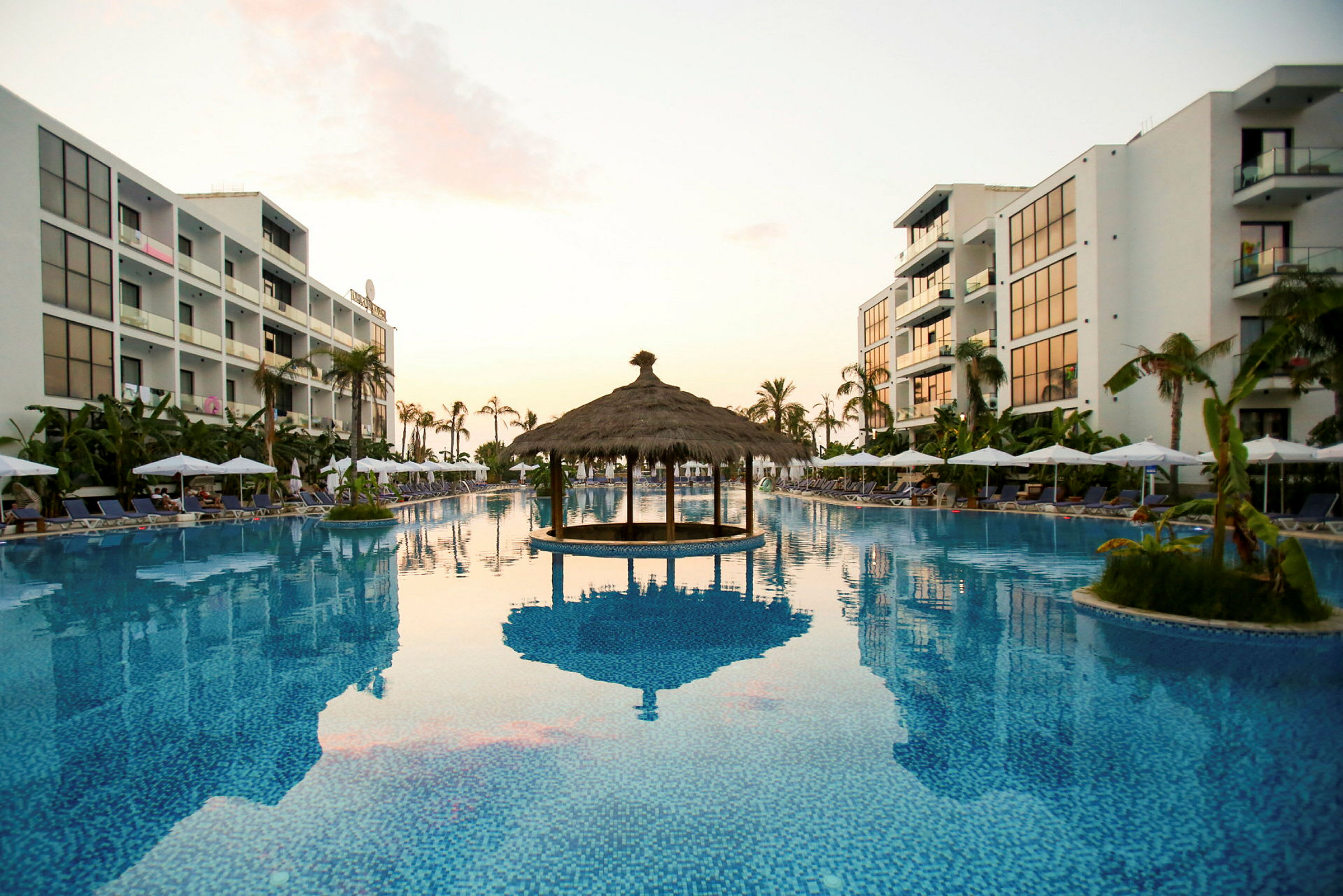 Ett hotellområde med en stor pool kantad av solstolar och parasoller, omgiven av palmer och moderna byggnader. (AI-genererad text)