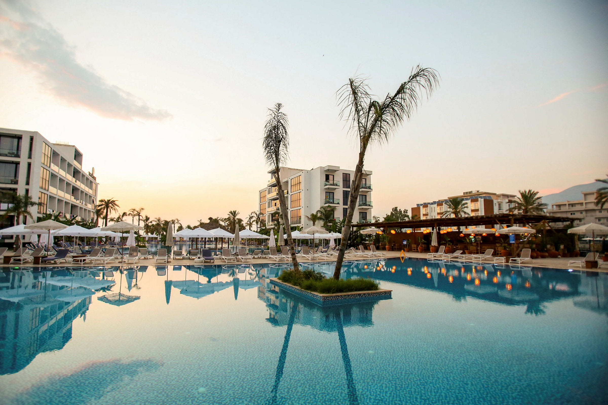 Bild av ett hotellområde med pool och omgivande solstolar och parasoller, samt byggnader i bakgrunden. (AI-genererad text)