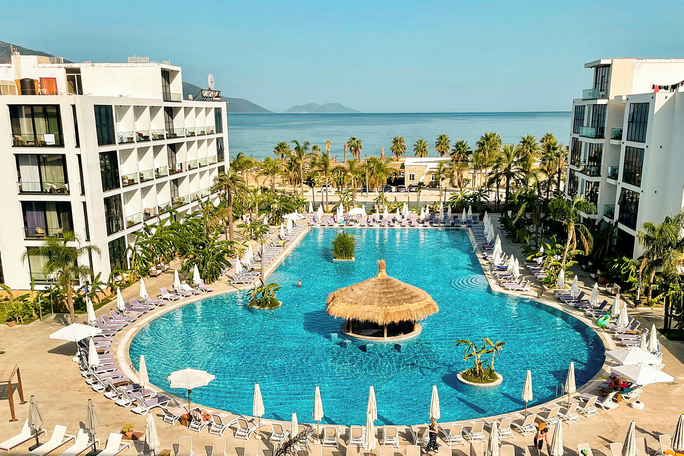 Hotellkomplex med stor pool omgiven av solstolar och parasoller, med havet i bakgrunden. (AI-genererad text)