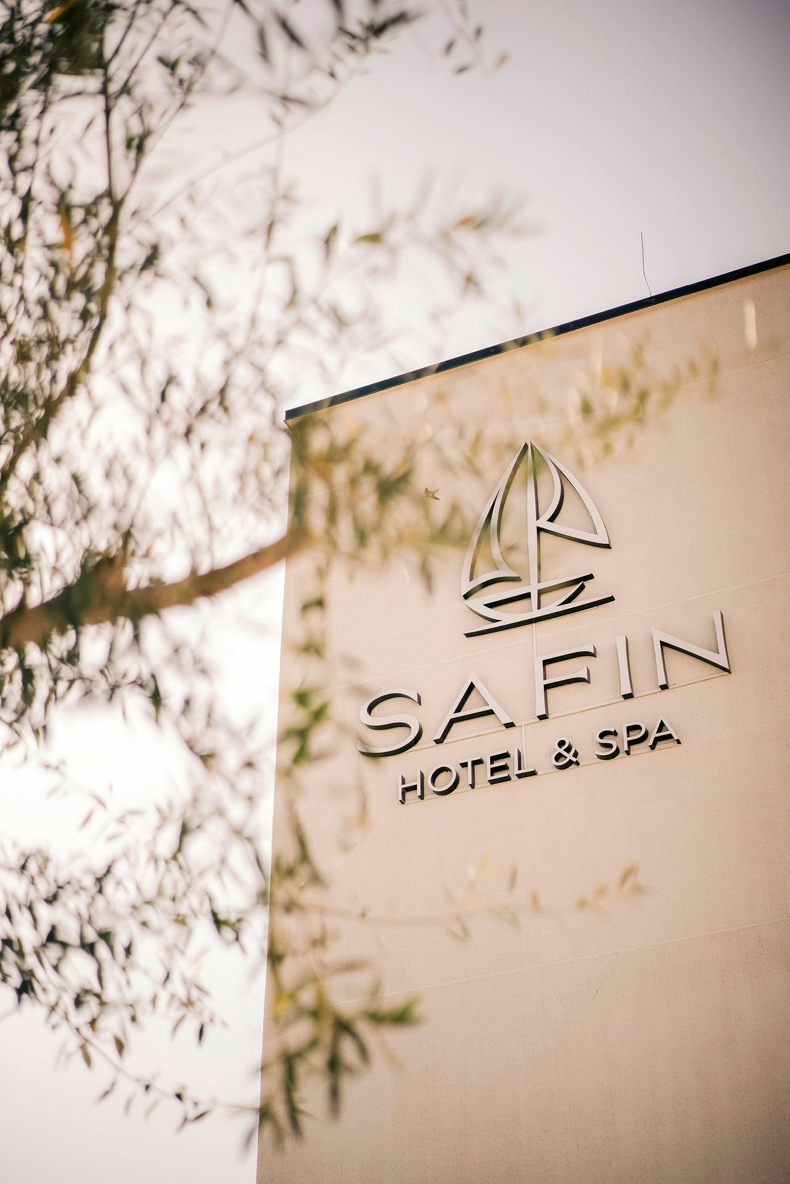 Byggnadsvägg med texten "SAFIN Hotel & Spa" och en logotyp som liknar en segelbåt. Delar av ett träd skymtar i förgrunden. (AI-genererad text)