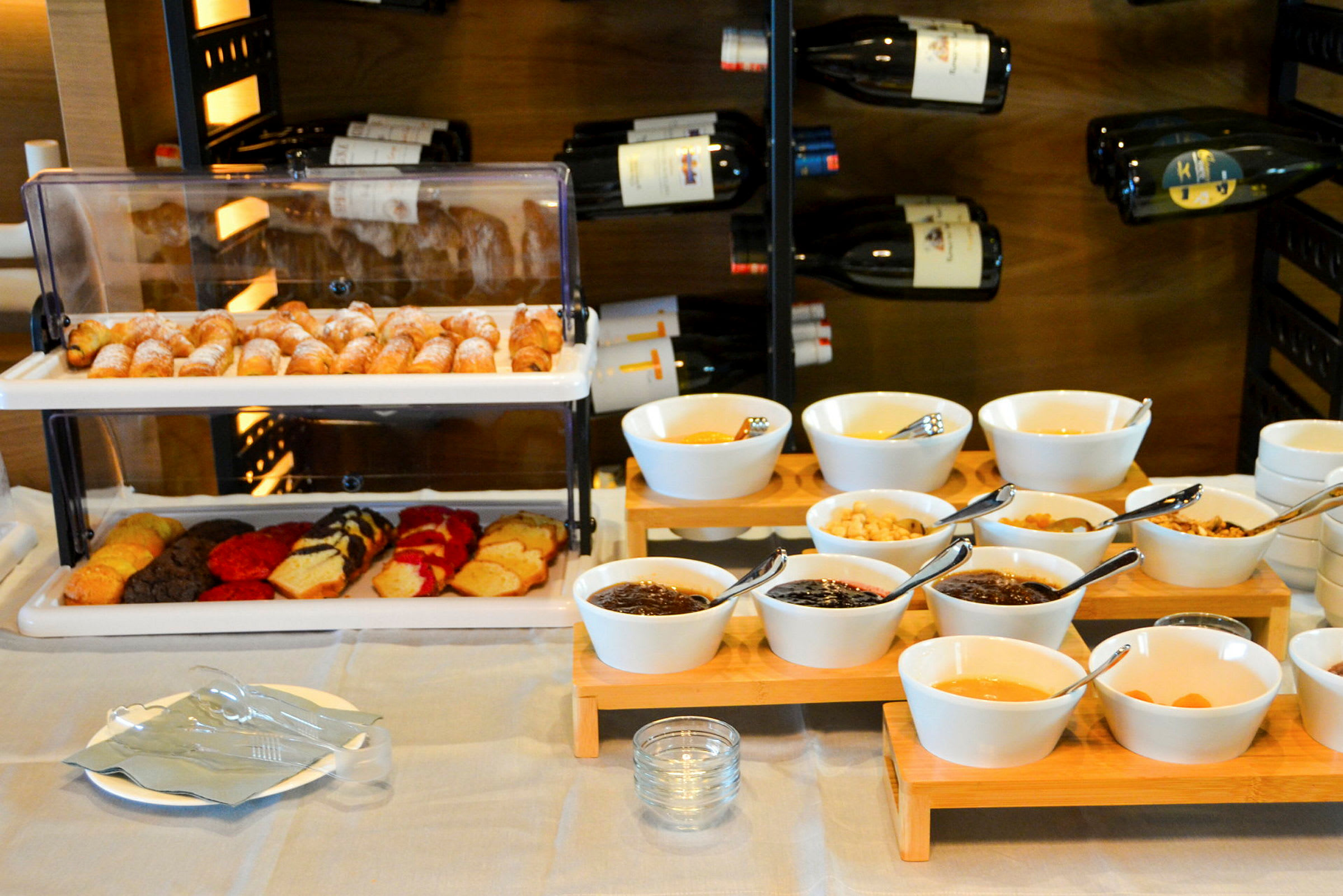 Ett frukostbord med croissanter, kakor och skålar med olika tillbehör framför en vinförvaring. (AI-genererad text)