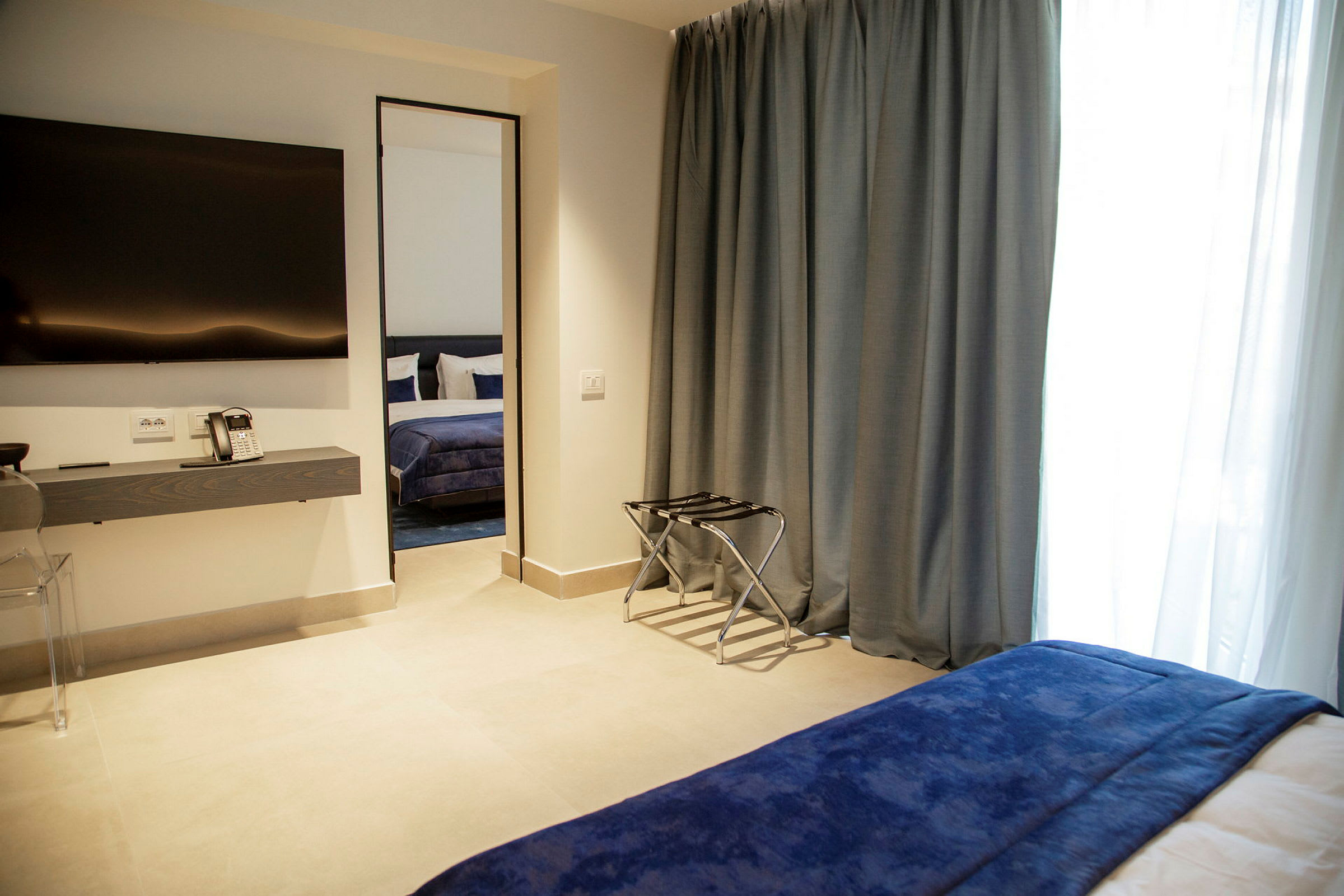Ett hotellrum med en TV på väggen, skrivbord med telefon, och en säng med blått överkast synlig genom en öppning. (AI-genererad text)