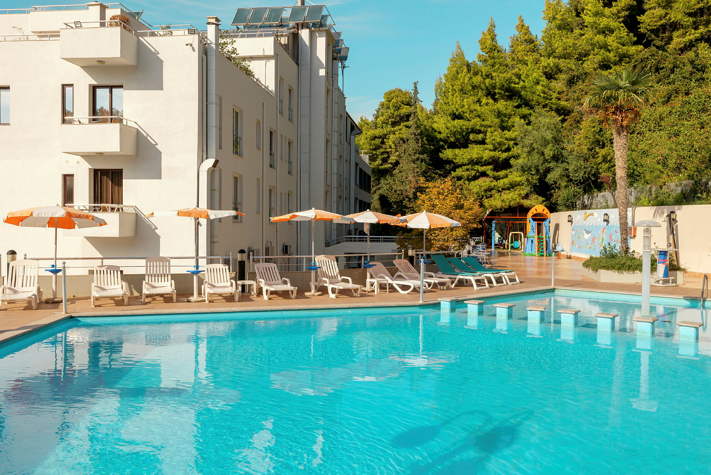 En utomhuspool omgiven av solstolar, parasoll och ett hotell i bakgrunden. (AI-genererad text)
