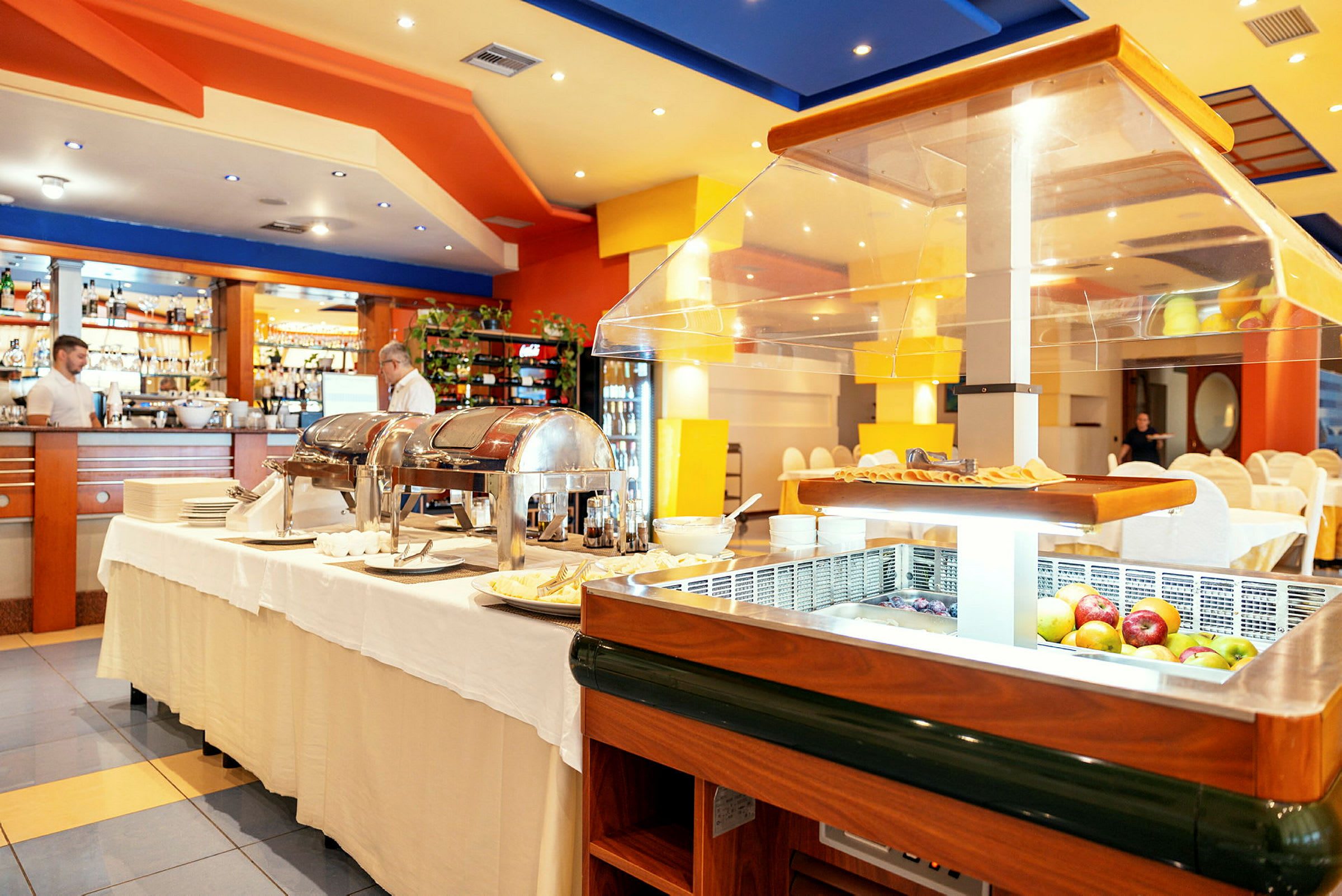 Bild av ett restauranginteriör med bufféutställning och glaspaneler på borden, samt dukade bord och stolar i bakgrunden. (AI-genererad text)