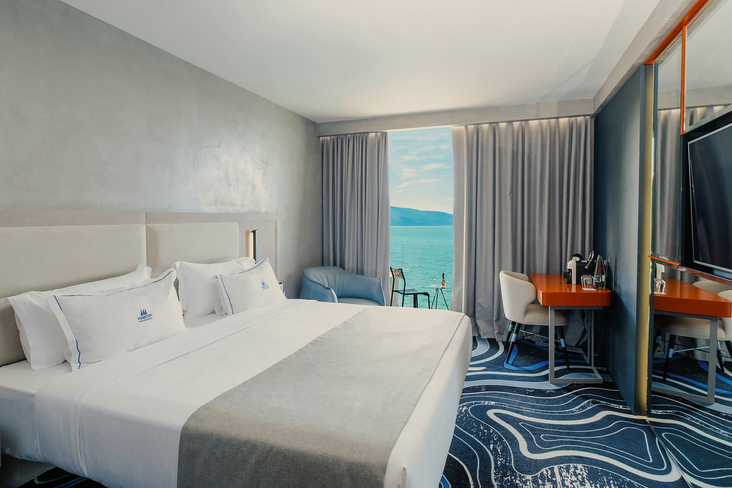 Ett hotellrum med en stor säng, en fåtölj och ett skrivbord. Utsikt över havet från ett fönster med gardiner. (AI-genererad text)