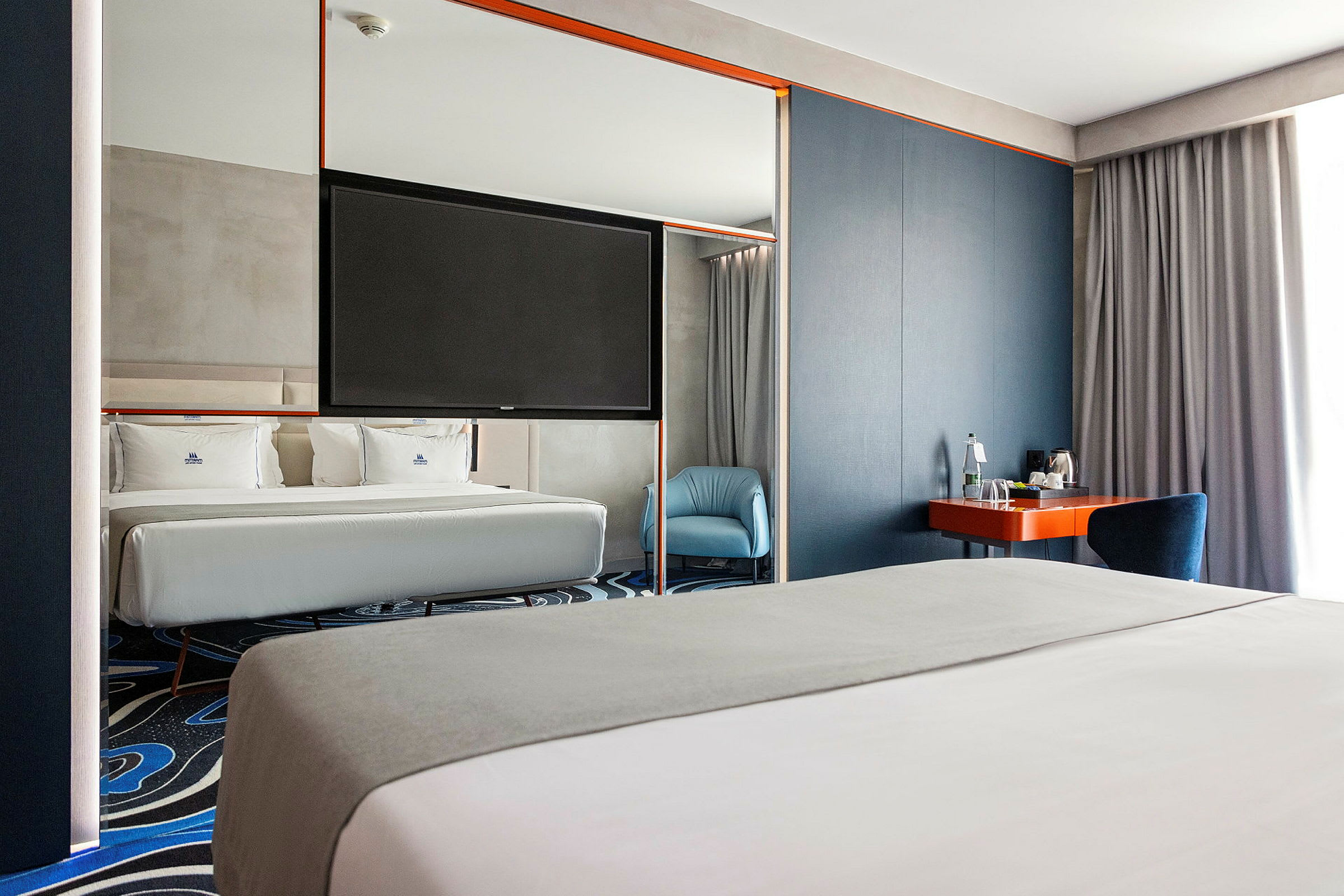 Ett hotellrum med en stor säng, en blå fåtölj och en arbetsyta med en spegel. (AI-genererad text)