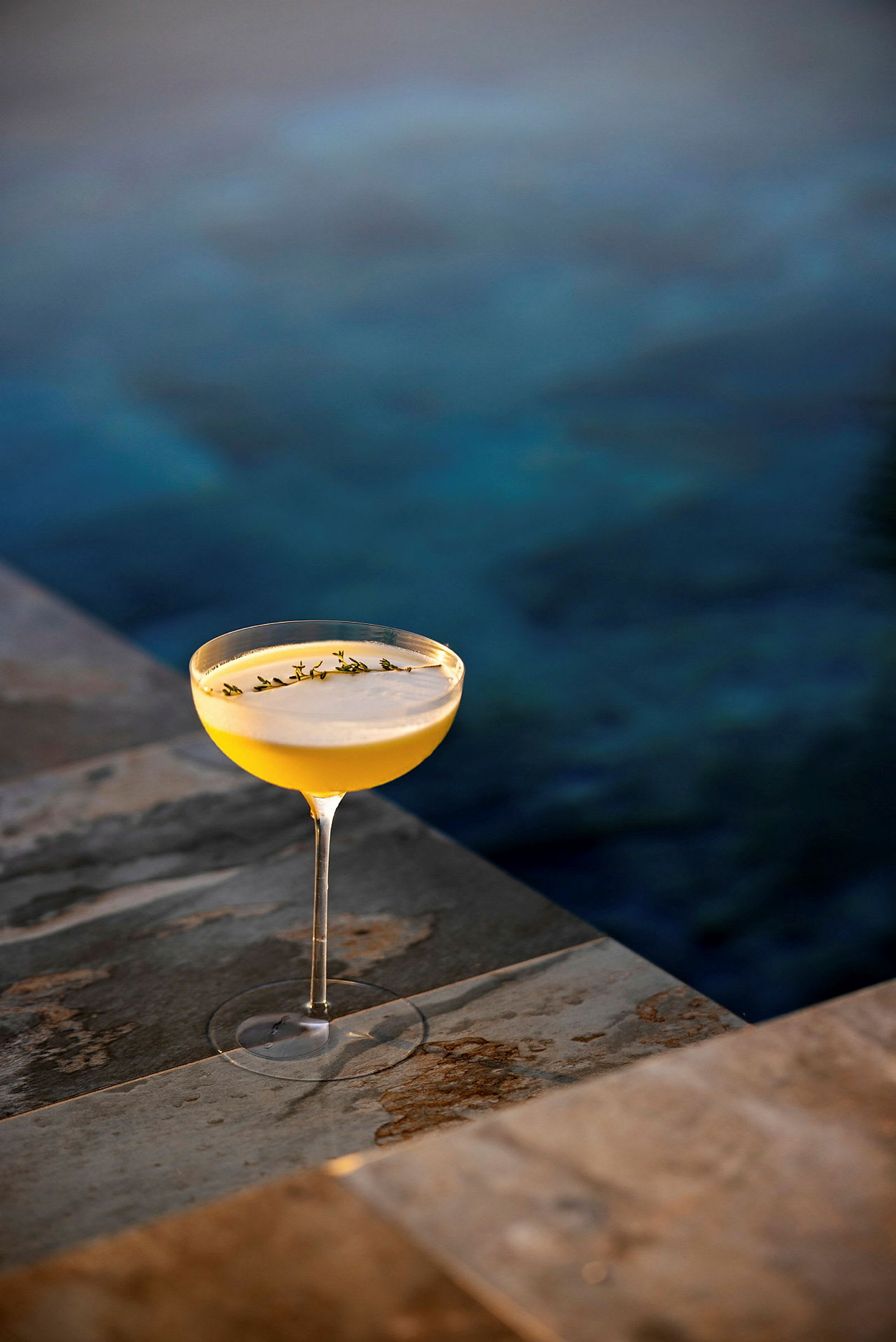 Ett glas med en gul drink och garnering står på en stenyta vid en pool. (AI-genererad text)