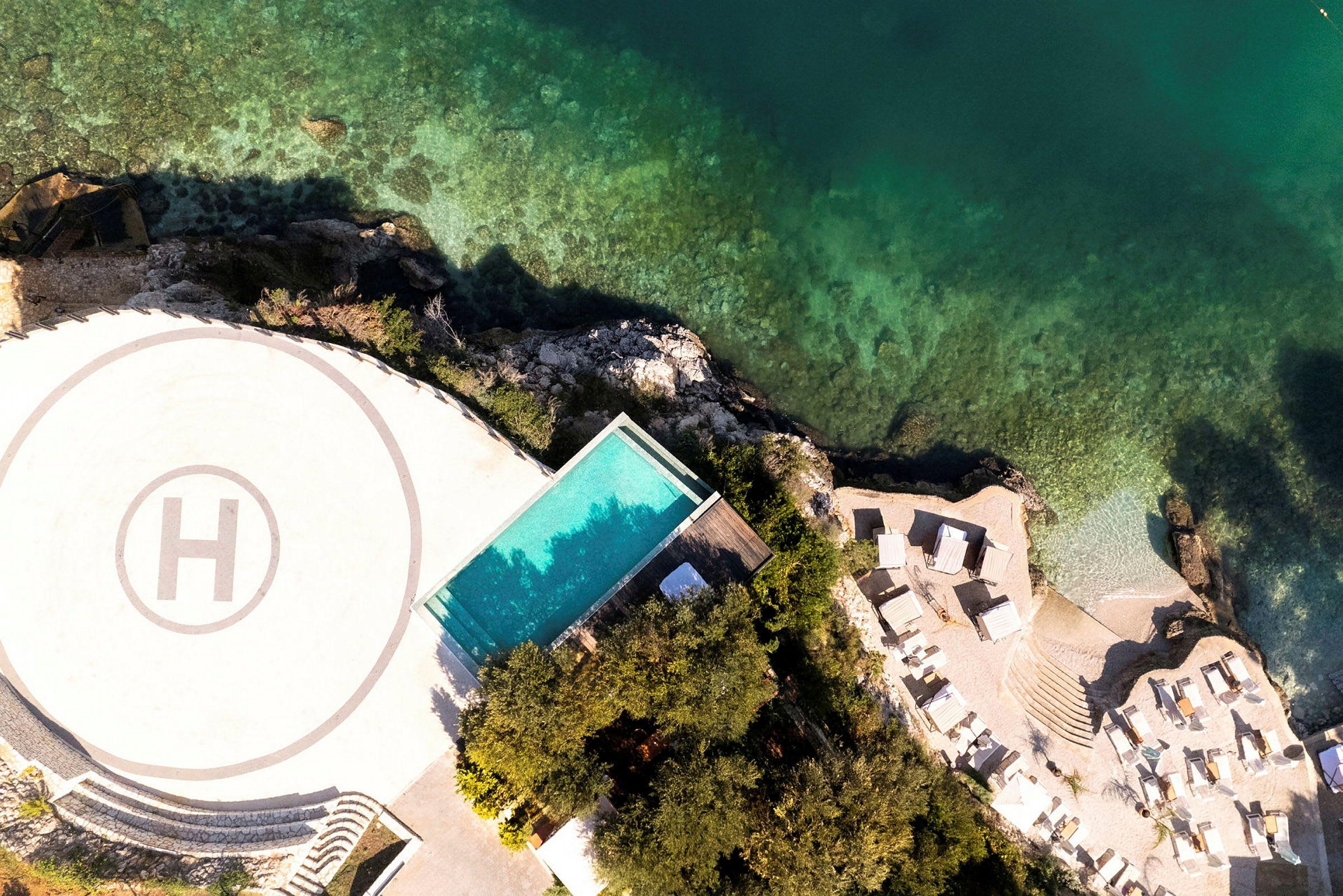 Flygbild av ett område med helikopterplatta, pool, solsängar, och kustlinje med klart vatten. (AI-genererad text)