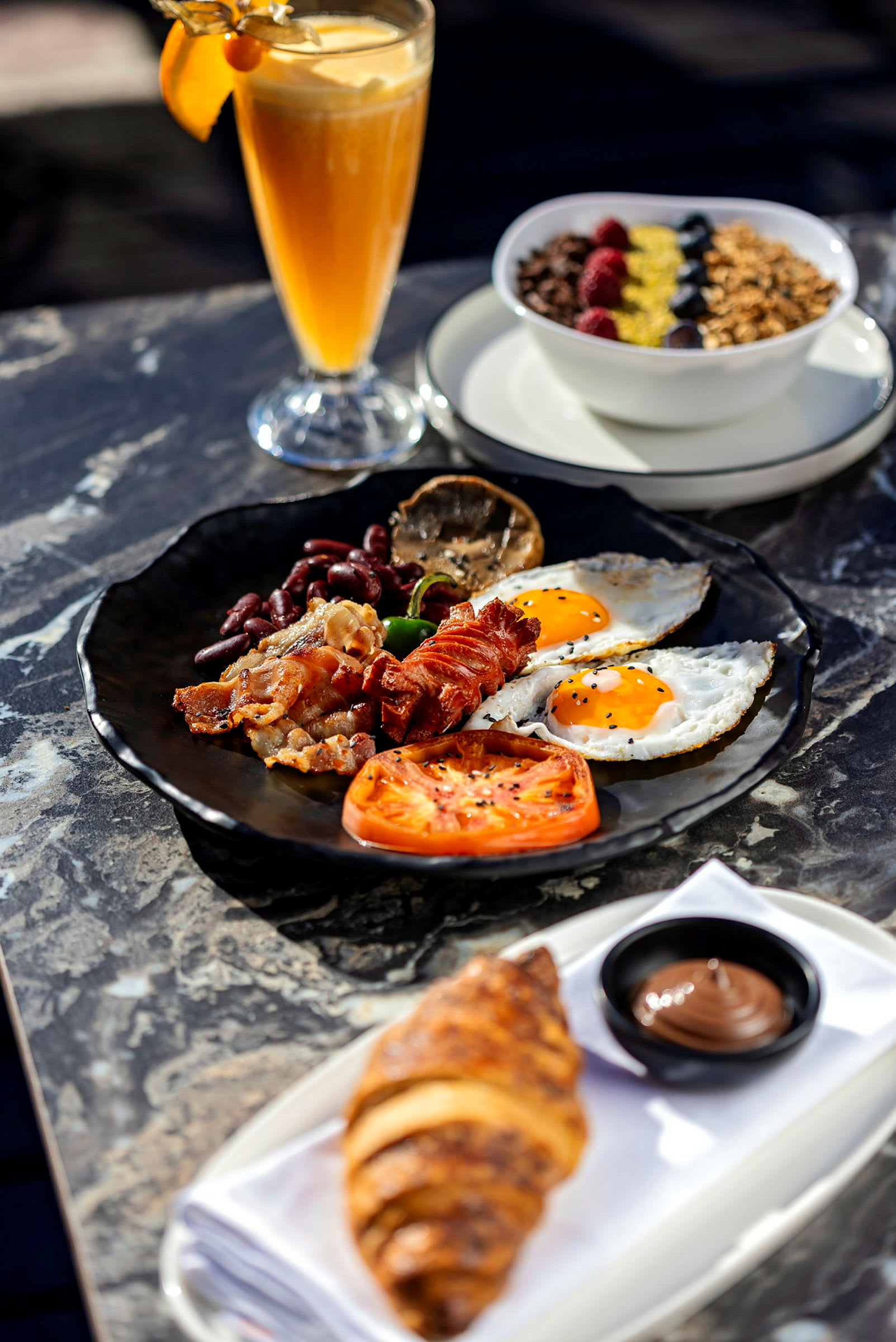 En tallrik med frukostmat bestående av bacon, ägg, tomat och grönsaker. En croissant med chokladkräm och en skål med flingor samt ett glas juice är också närvarande. (AI-genererad text)