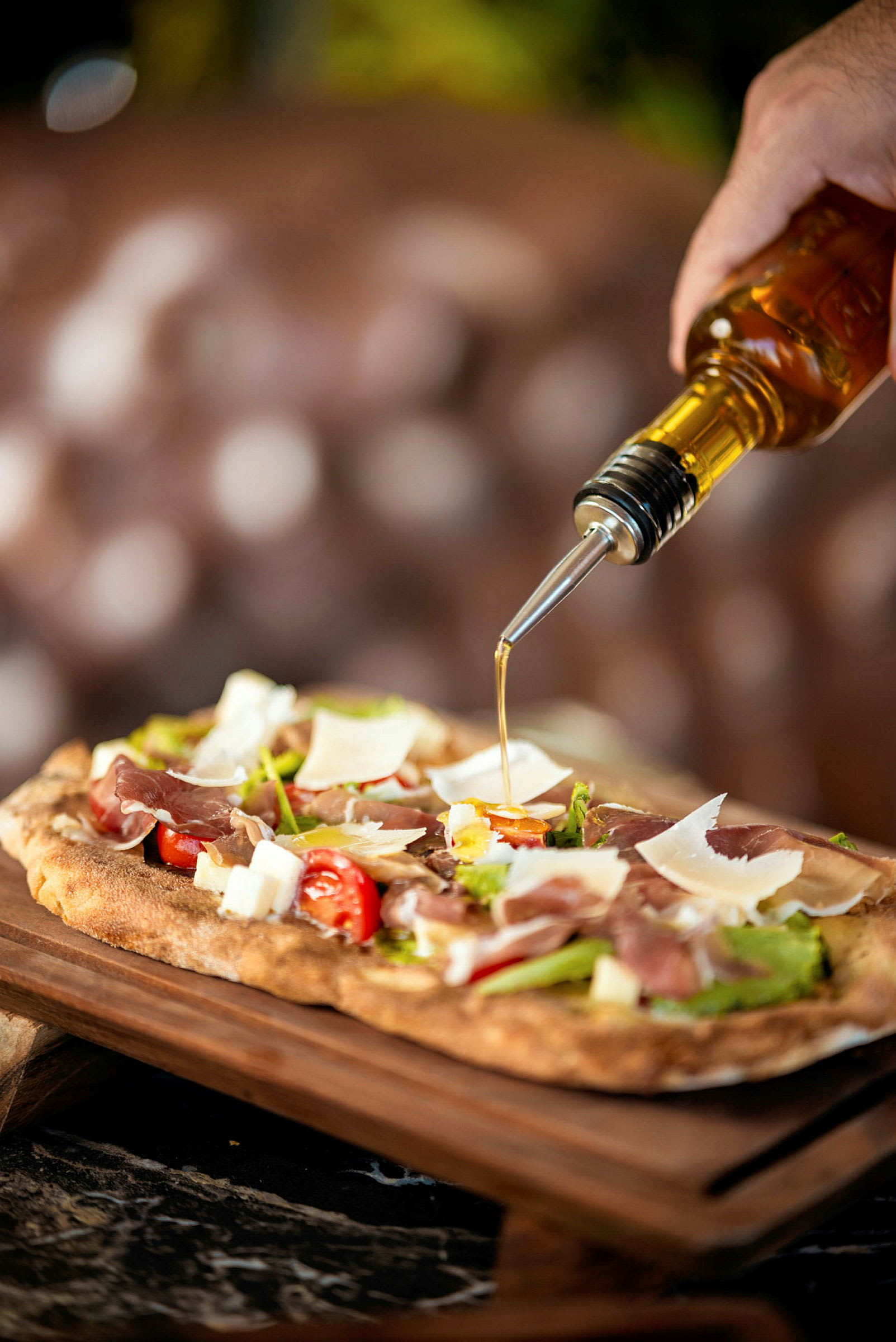 En hand häller olivolja från en flaska över en pizza med skinka, tomater och parmesanost. (AI-genererad text)