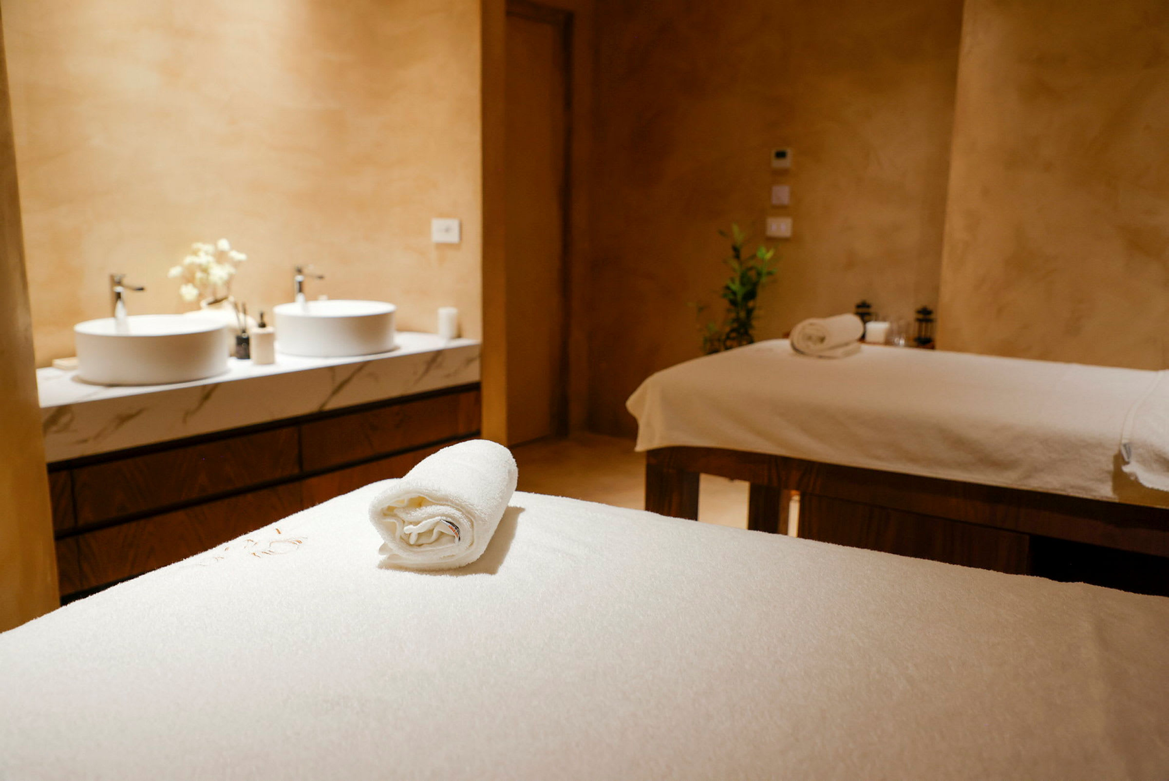 Ett spa-rum med två behandlingsbänkar täckta med vita handdukar och två handfat med speglar i bakgrunden. (AI-genererad text)