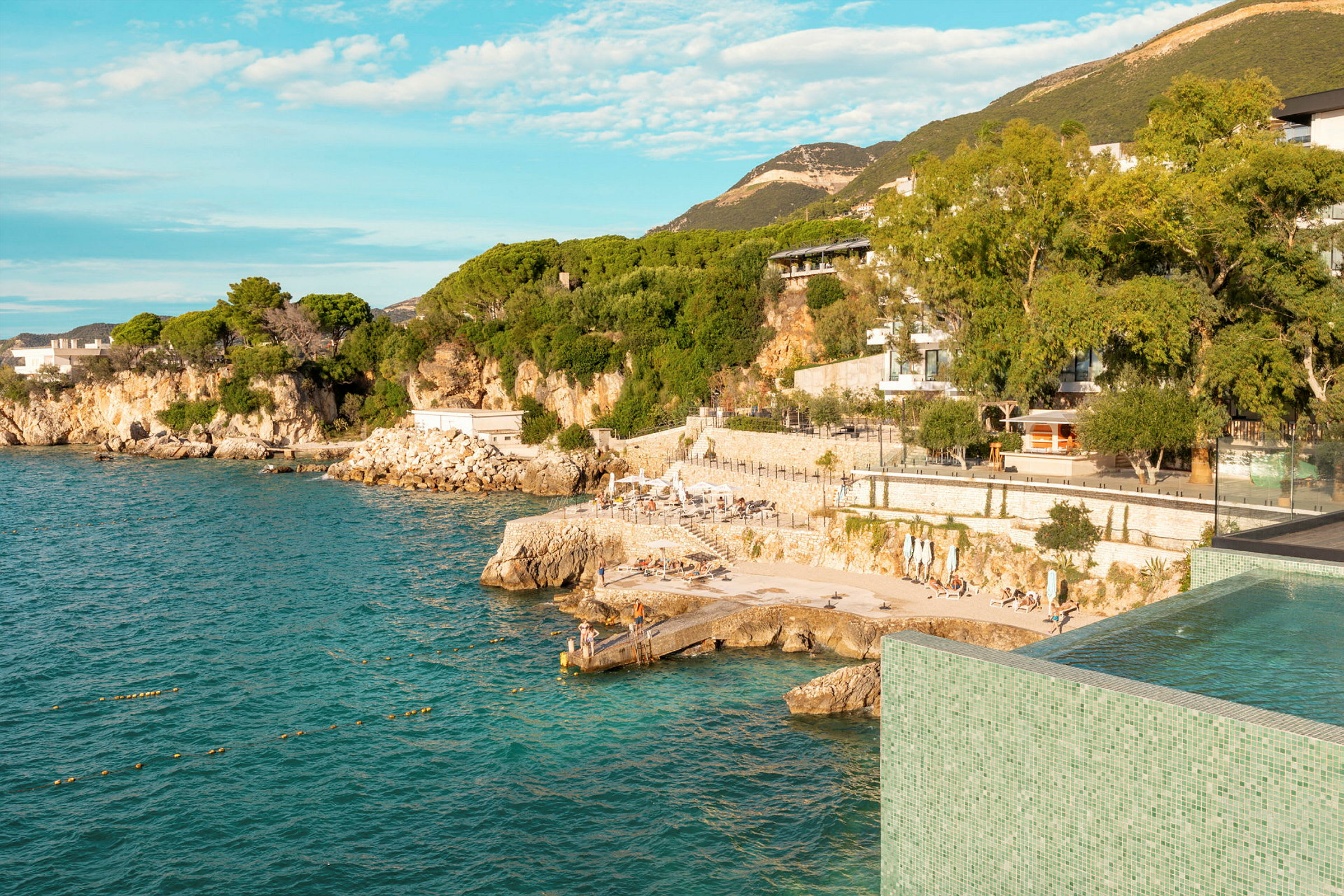 En kustlinje med gröna träd och berg i bakgrunden. Kustklippor och en upphöjd pool med utsikt över havet syns. (AI-genererad text)