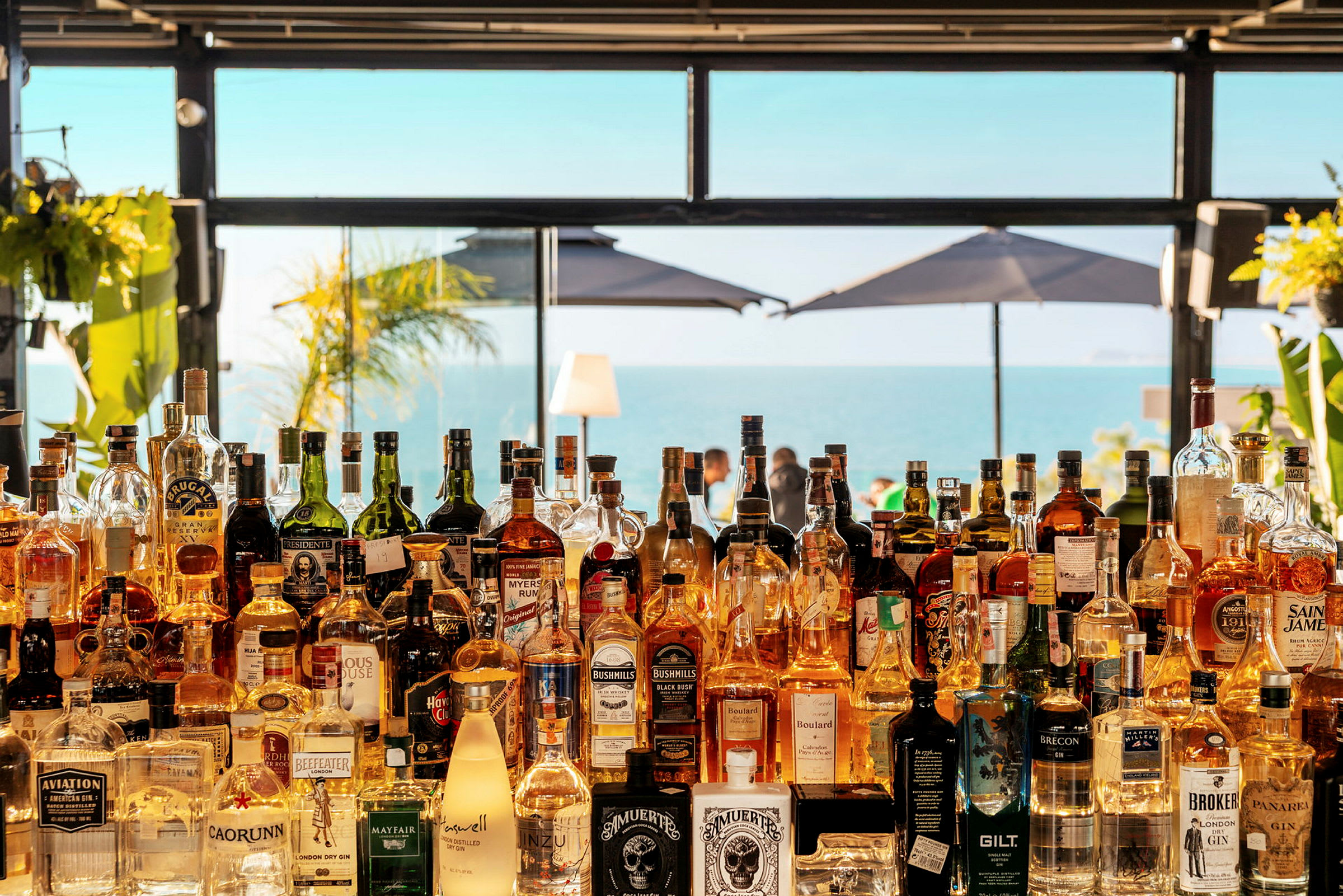 Utsikt över en strandbar med flaskor på en hylla och havet i bakgrunden. (AI-genererad text)