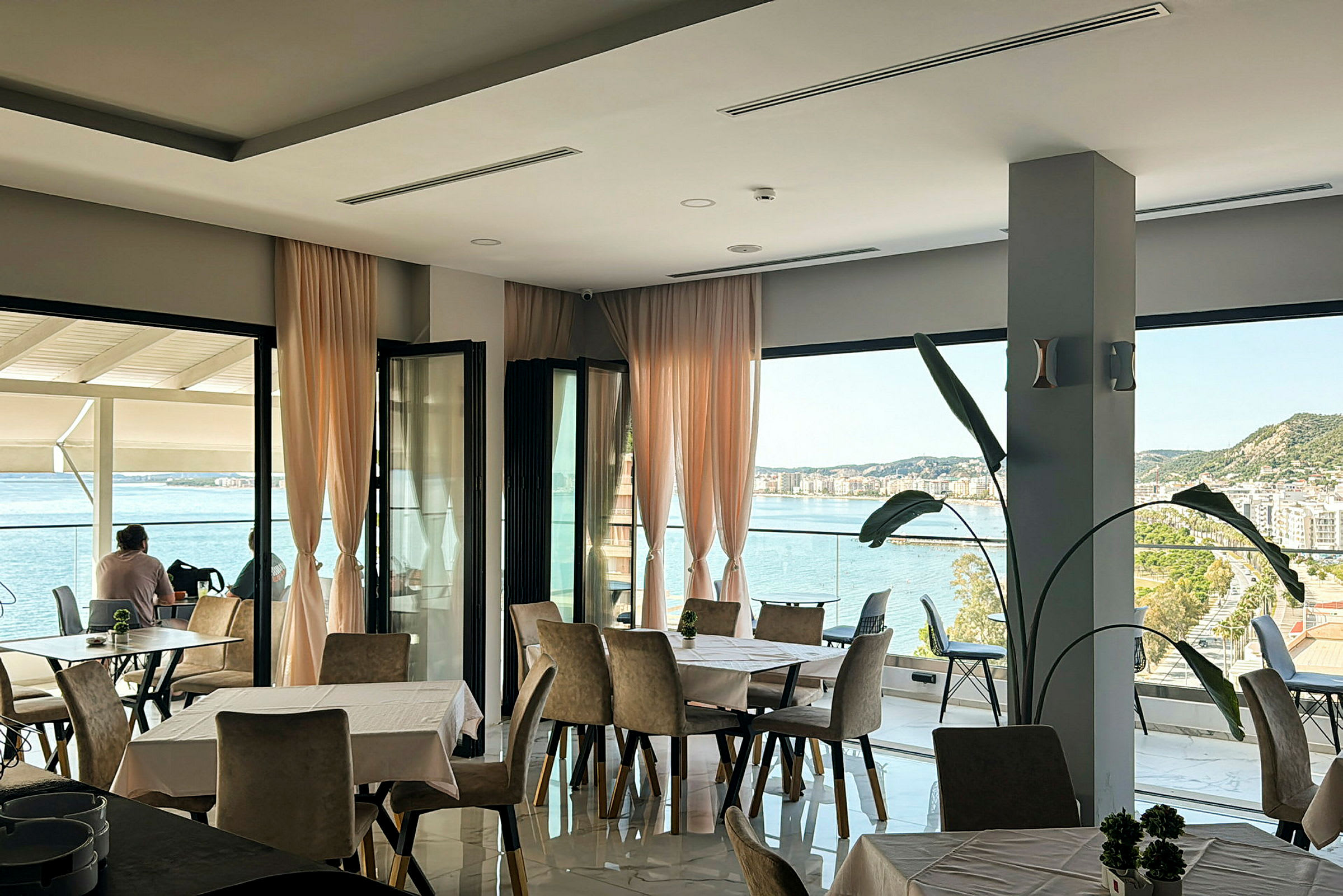 Ett restaurangområde med stora fönster och utsikt över havet och en stad. (AI-genererad text)