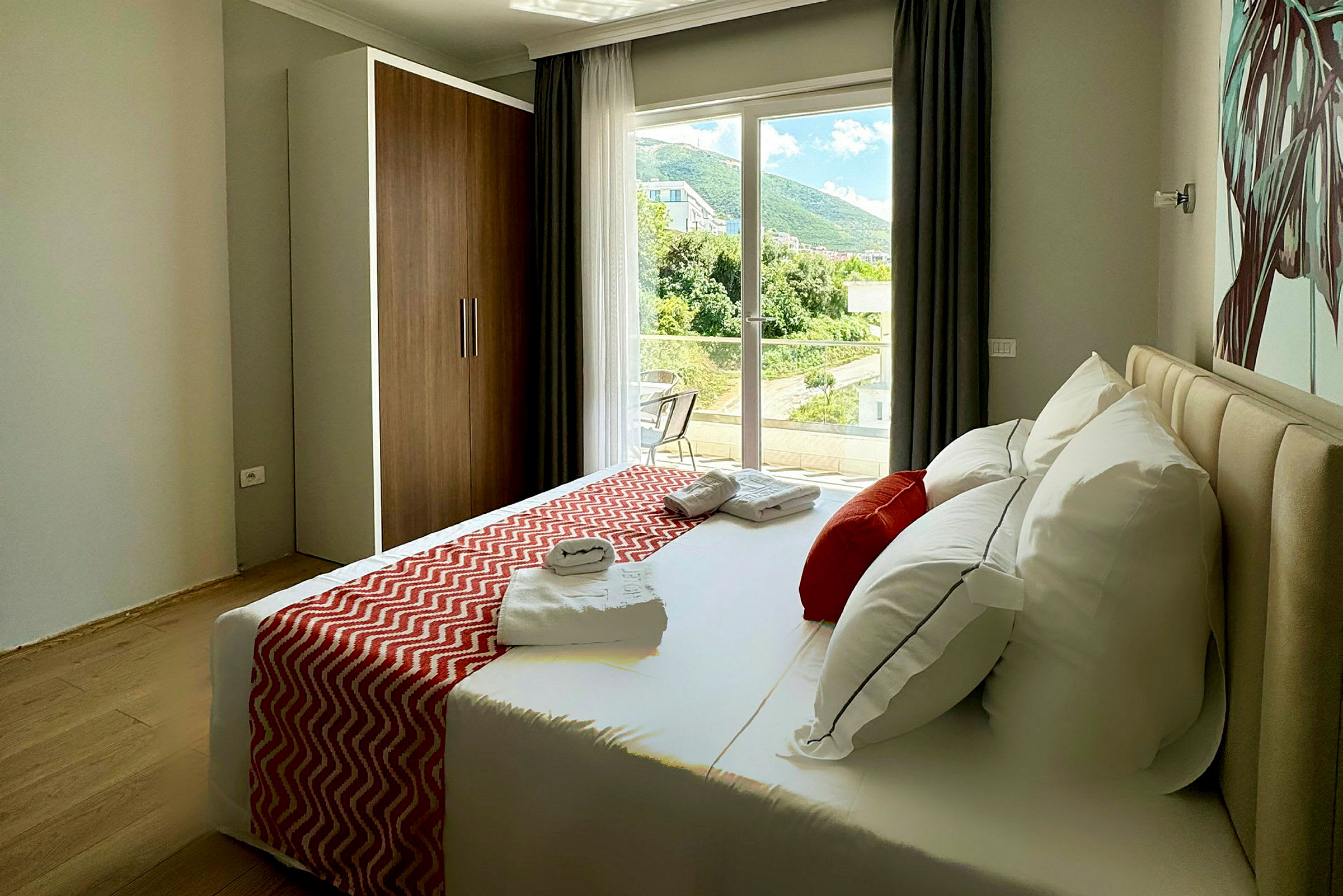Ett hotellrum med en säng, dekorerad med röda kuddar och sängöverkast, samt utsikt mot ett grönskande landskap från fönstret. (AI-genererad text)