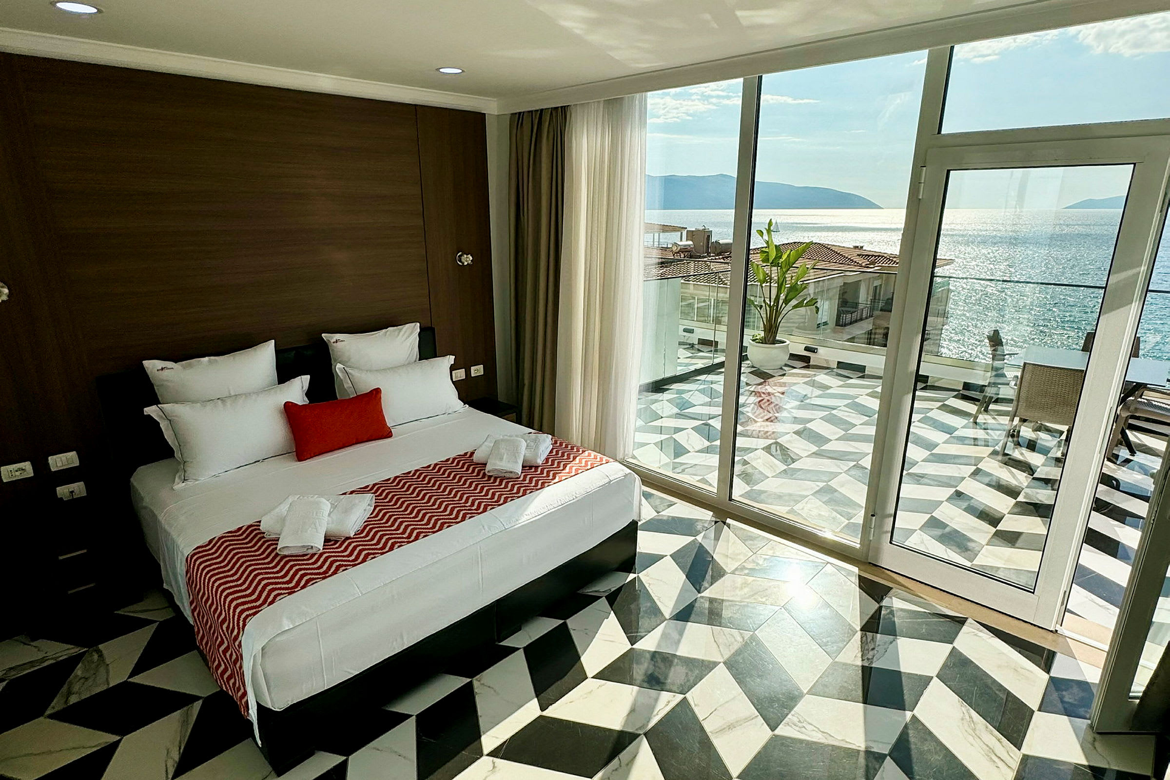 Ett hotellrum med en bred säng, modern inredning och en stor glasdörr som leder till en terrass med havsutsikt. (AI-genererad text)