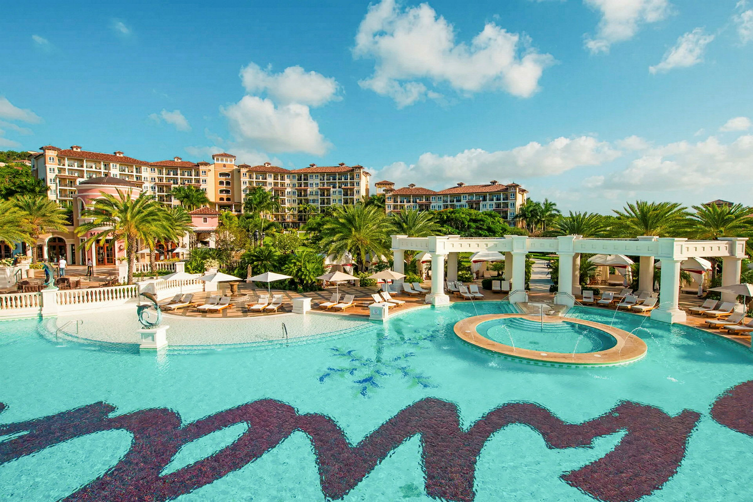 Bild av ett hotellkomplex med poolområde, parasoller och gröna palmer. (AI-genererad text)
