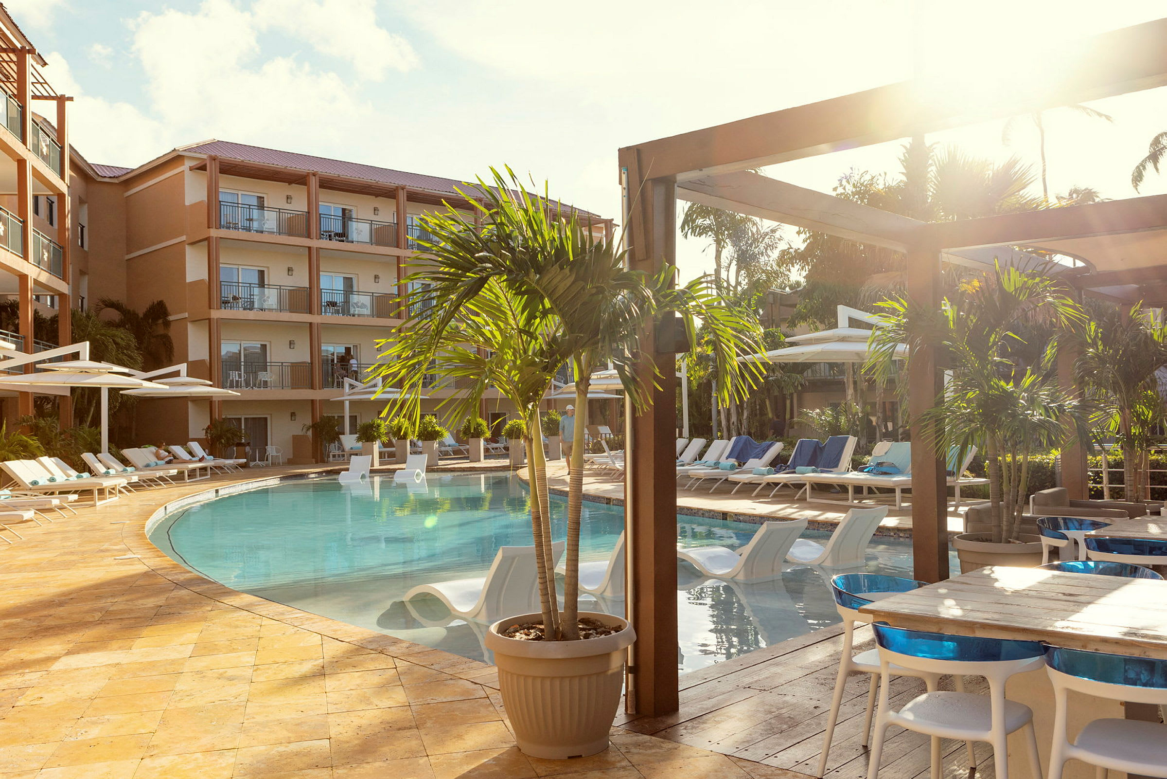 Bild av ett hotellområde med en pool omgiven av solstolar och parasoller. Framför finns bord och stolar placerade vid en träterrass. (AI-genererad text)