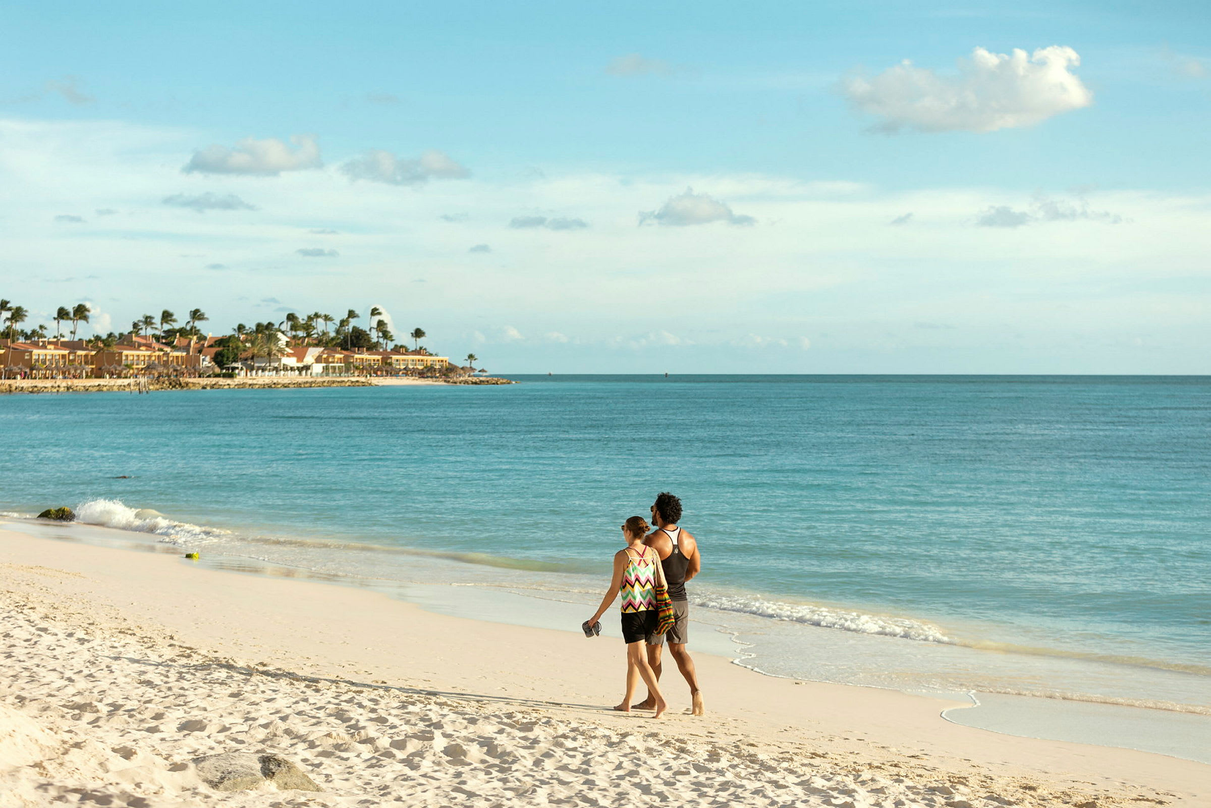 Ett par promenerar längs en sandstrand med utsikt över havet och byggnader längs strandlinjen. (AI-genererad text)