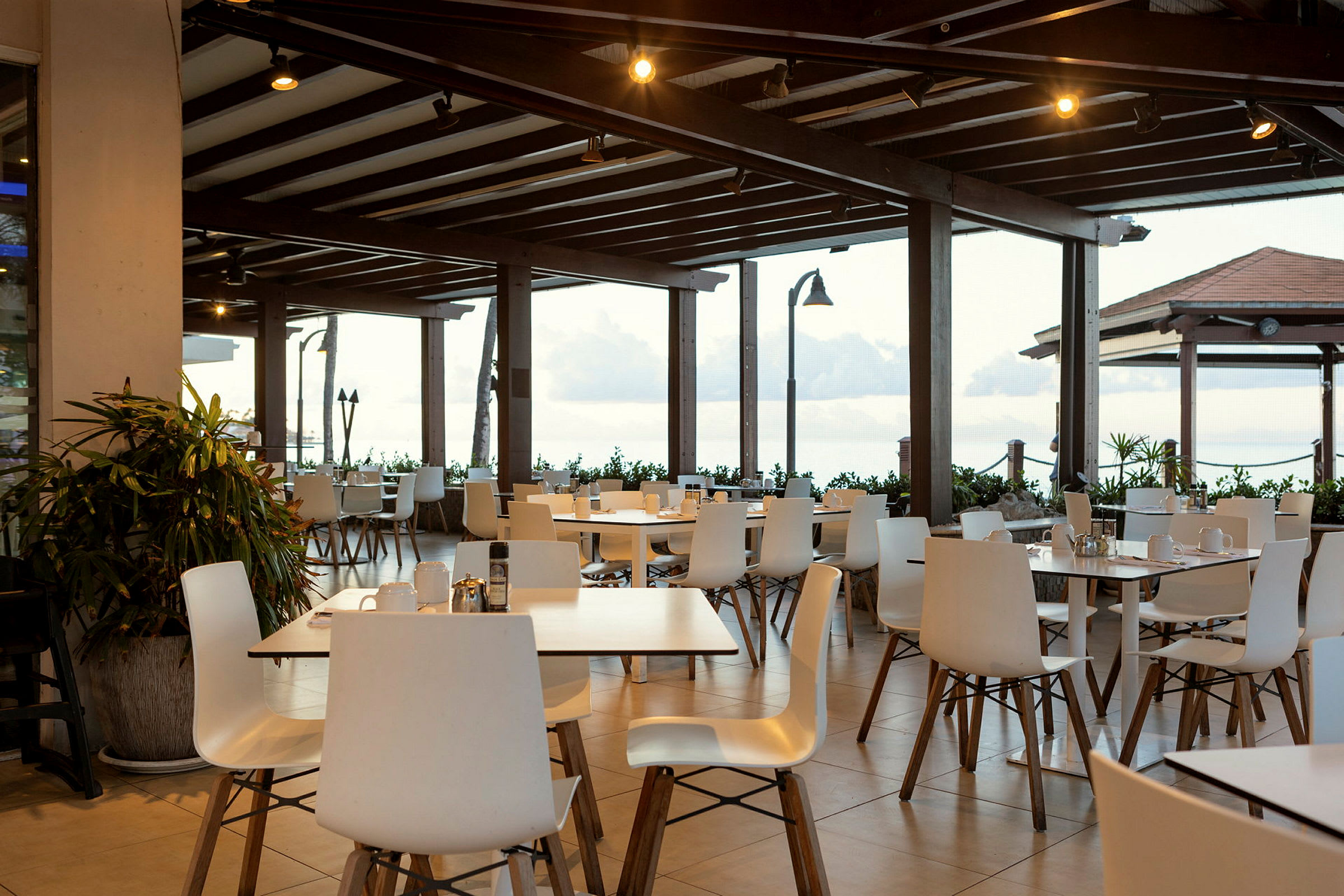 En restaurangterrass med vita stolar och bord under ett trätak. Utsikt över havet bakom. (AI-genererad text)
