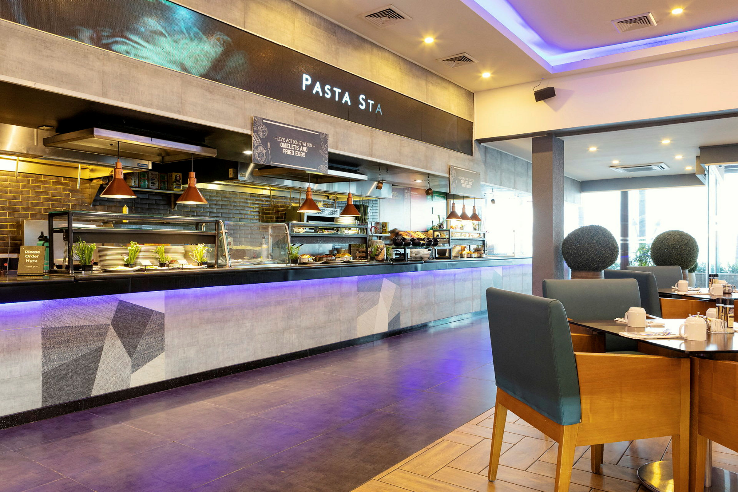 En restaurang med en öppen serveringsdisk och sittplatser. Bord med stolar syns i förgrunden, och bakom serveringsdisken visas en skylt med texten 'Pasta Sta'. (AI-genererad text)