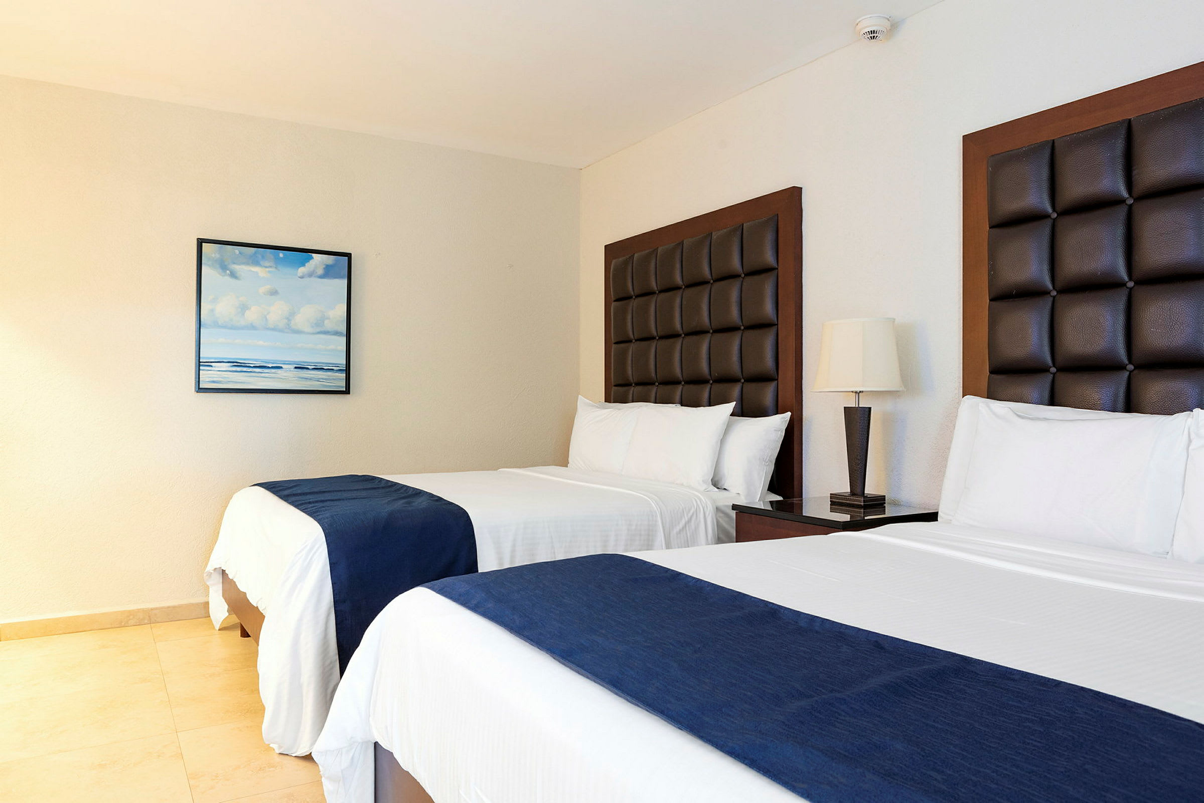 Två enkelsängar med mörkbruna sänggavlar och vita sängkläder med blå plädar, placerade i ett hotellrum. På väggen hänger en tavla med motiv av hav och moln. (AI-genererad text)