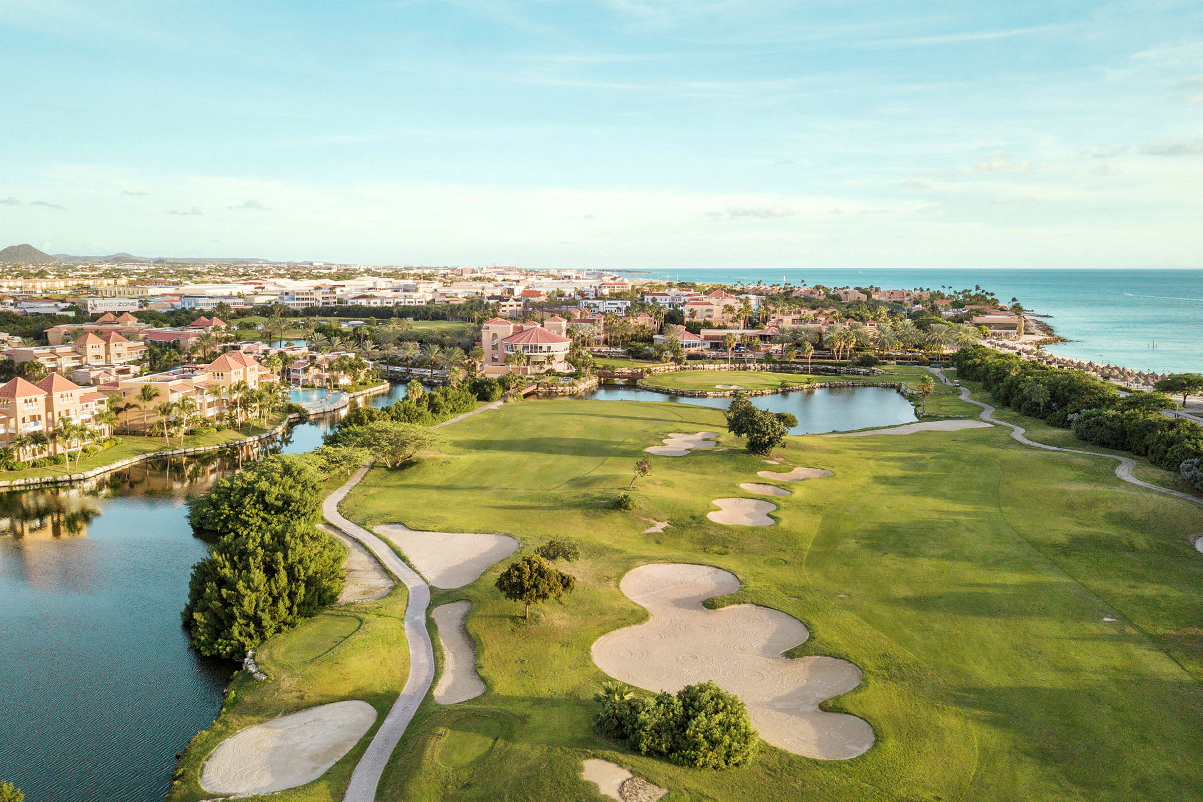 Golfbana med sandbunkrar och gröna gräsytor i närheten av vattendrag och strand. Bebyggelse i bakgrunden. (AI-genererad text)