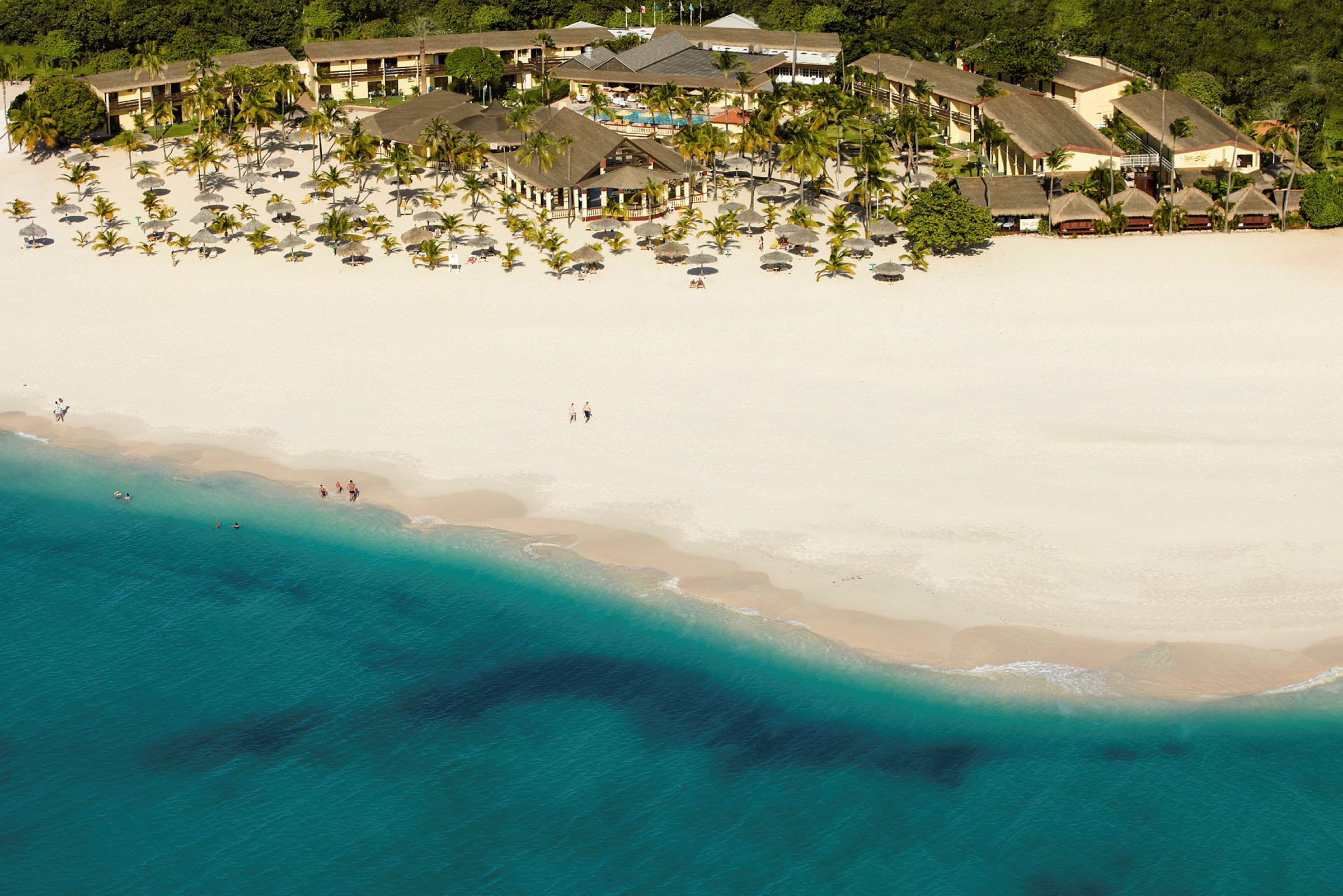 Flygfoto av en vit sandstrand med blått hav framför ett hotellområde omgärdat av palmer. (AI-genererad text)