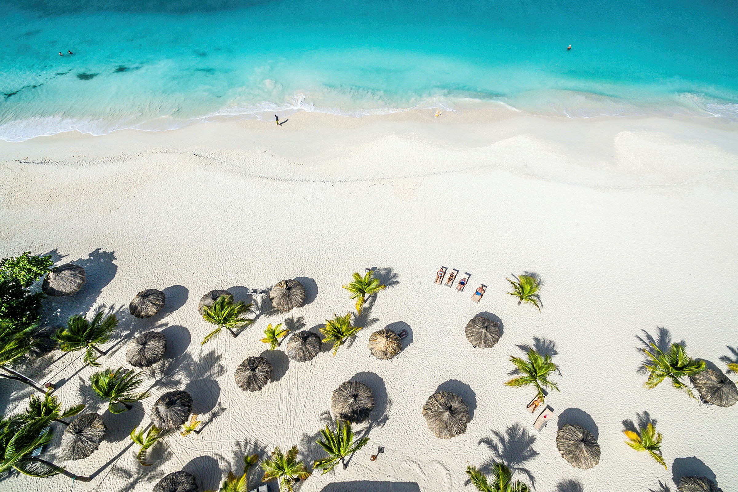Flygfoto av en vit sandstrand med turkost vatten. Stranden är dekorerad med parasoller och omgiven av palmer. (AI-genererad text)