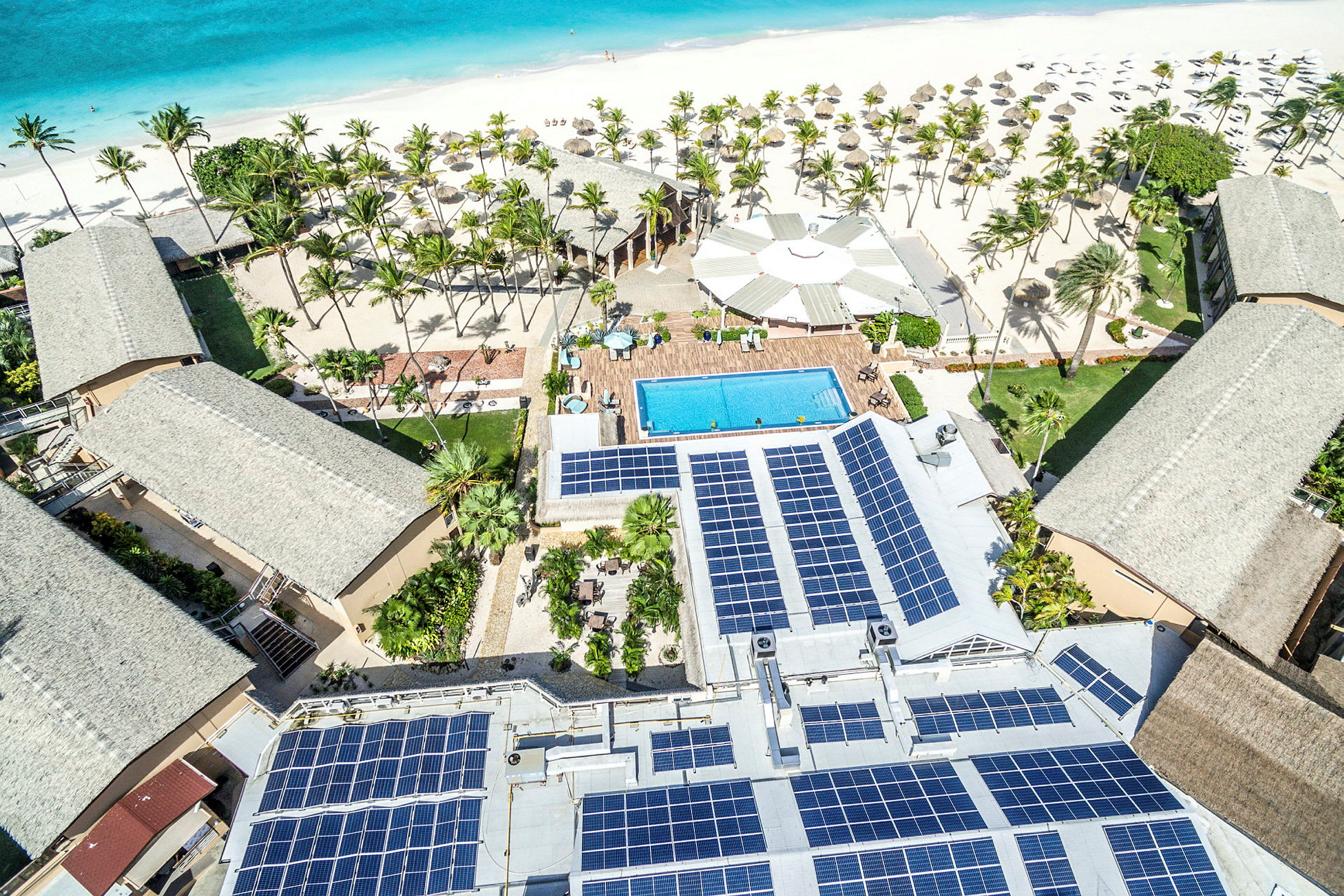 Flygfoto av ett semesterdestination nära en vit sandstrand med palmer, en swimmingpool, och byggnader med solpaneler. (AI-genererad text)