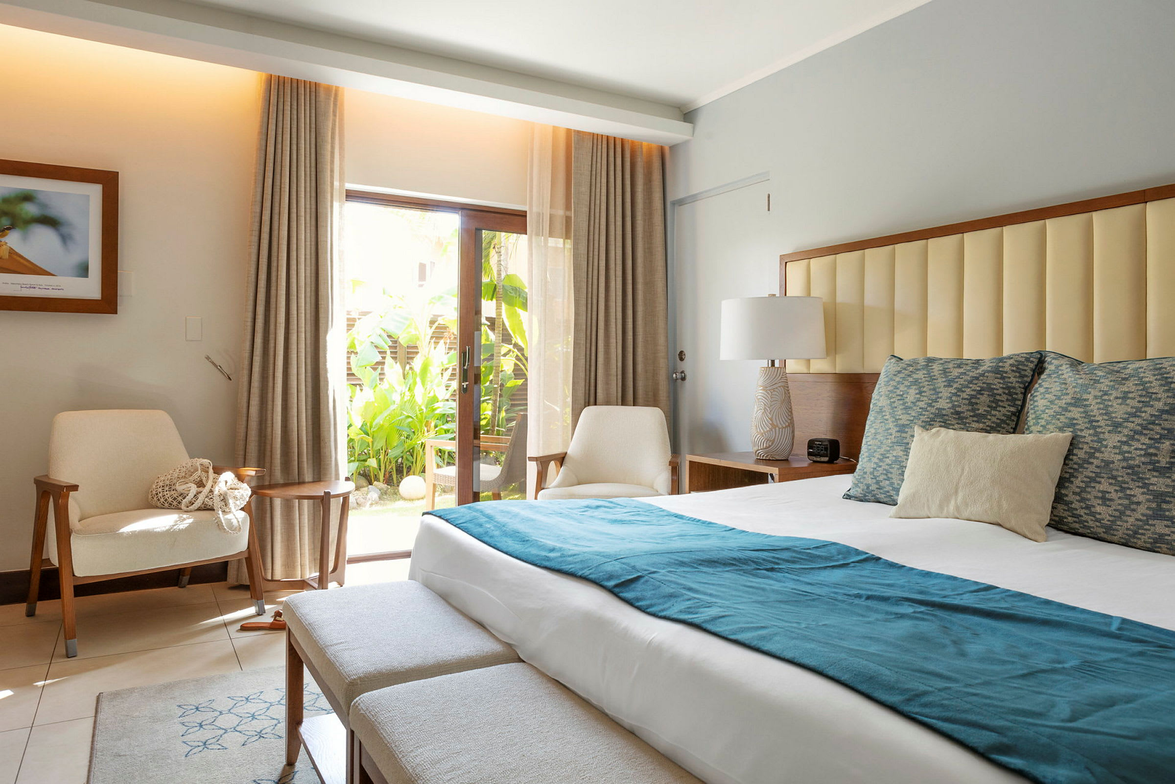 Ett ljust hotellrum med en dubbelsäng klädd i vita lakan och blå detaljer, två fåtöljer, och en glasdörr som leder till en tropisk trädgård. (AI-genererad text)