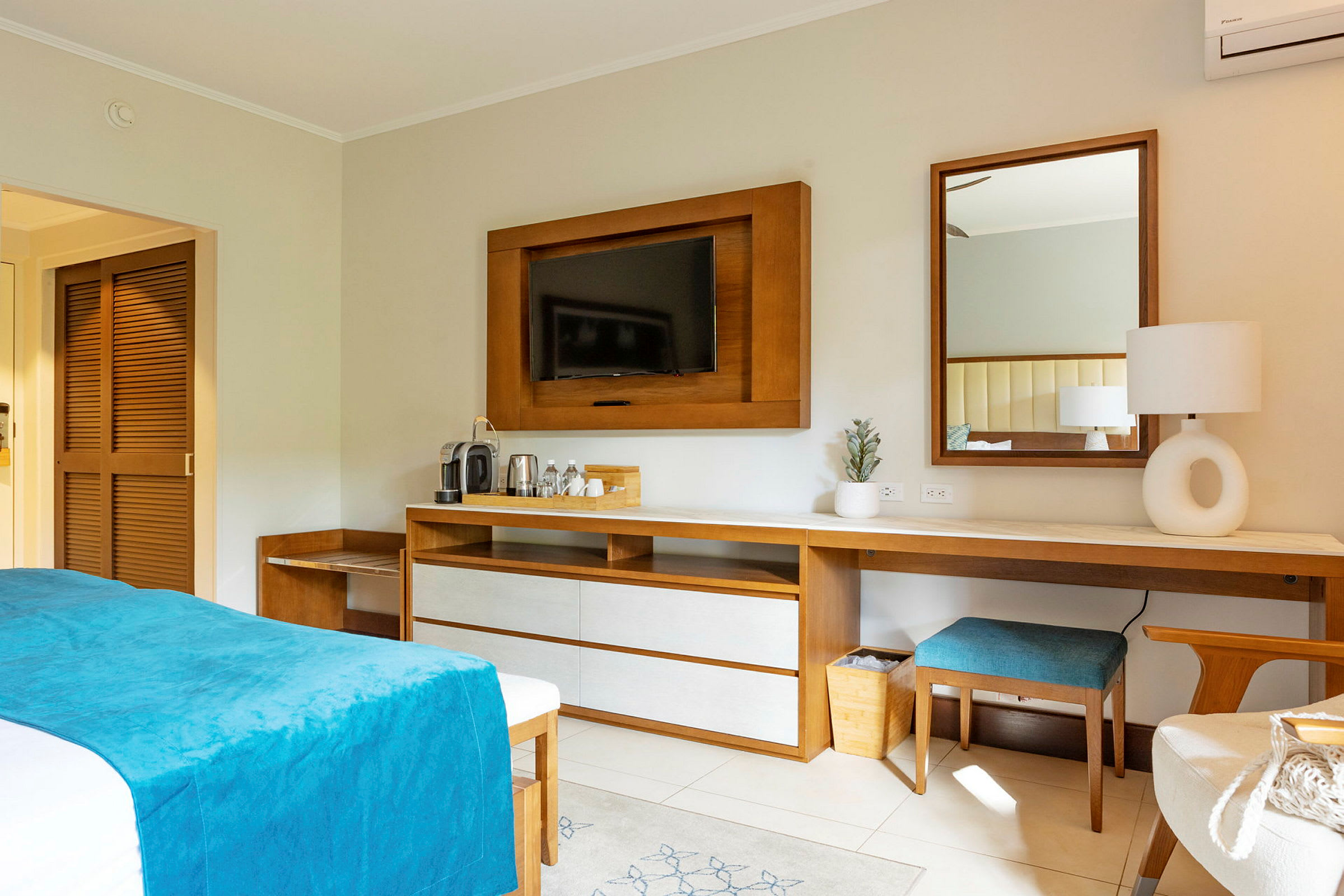 Ett hotellrum med en säng med blå överkast, träinredning, en TV på väggen och ett skrivbord med spegel och lampa. (AI-genererad text)