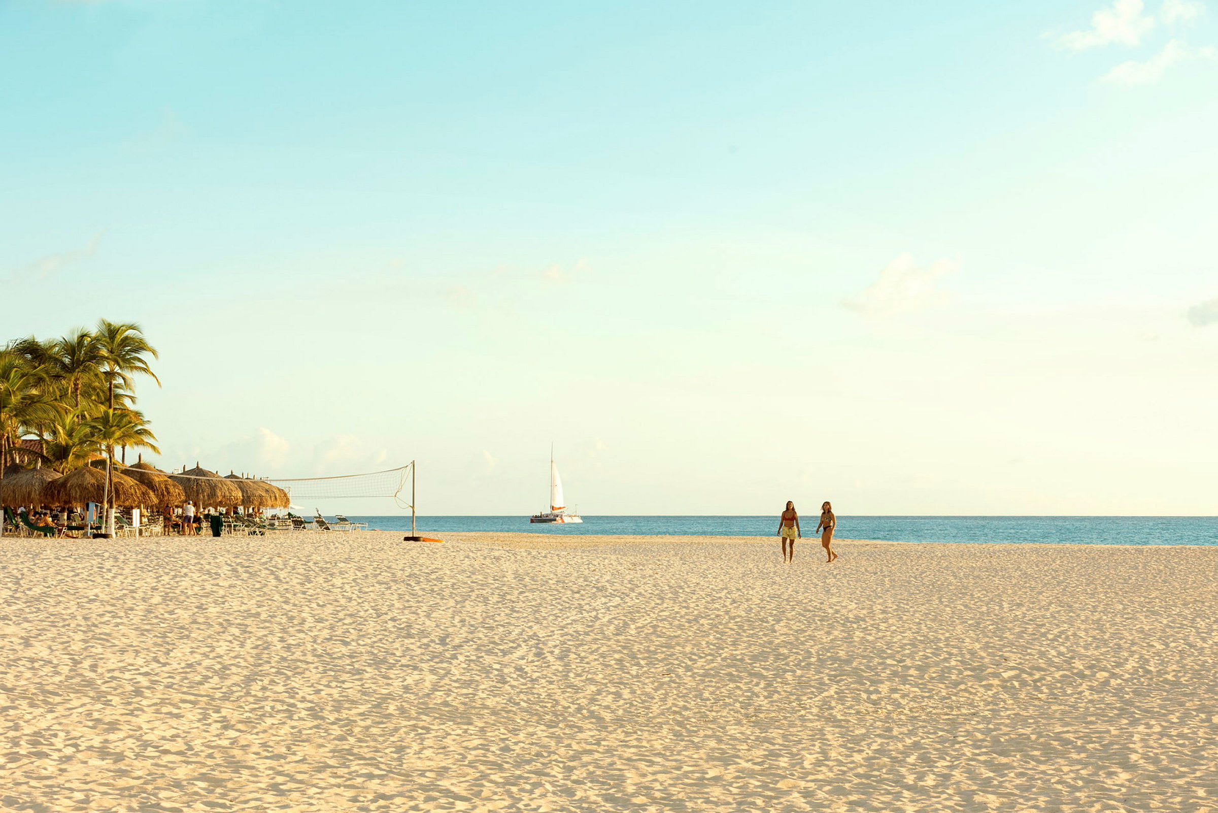 En sandstrand med palmer och parasoll till vänster, två personer som går på stranden och en segelbåt på havet. (AI-genererad text)
