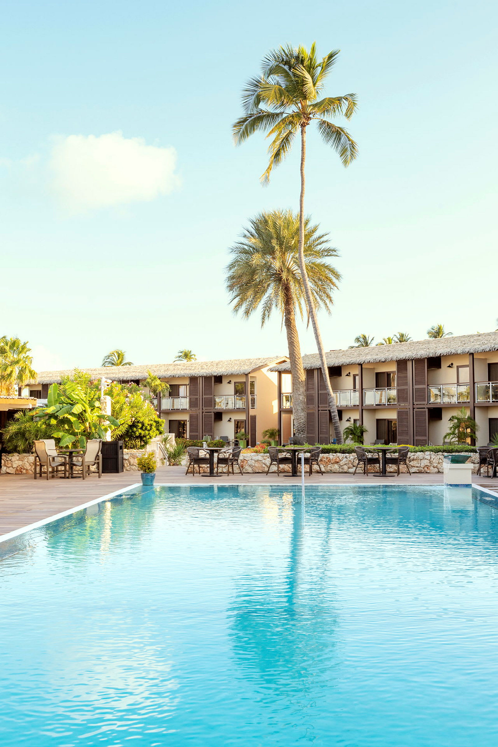 En pool omgiven av bord och stolar framför ett hotell med palmer. (AI-genererad text)