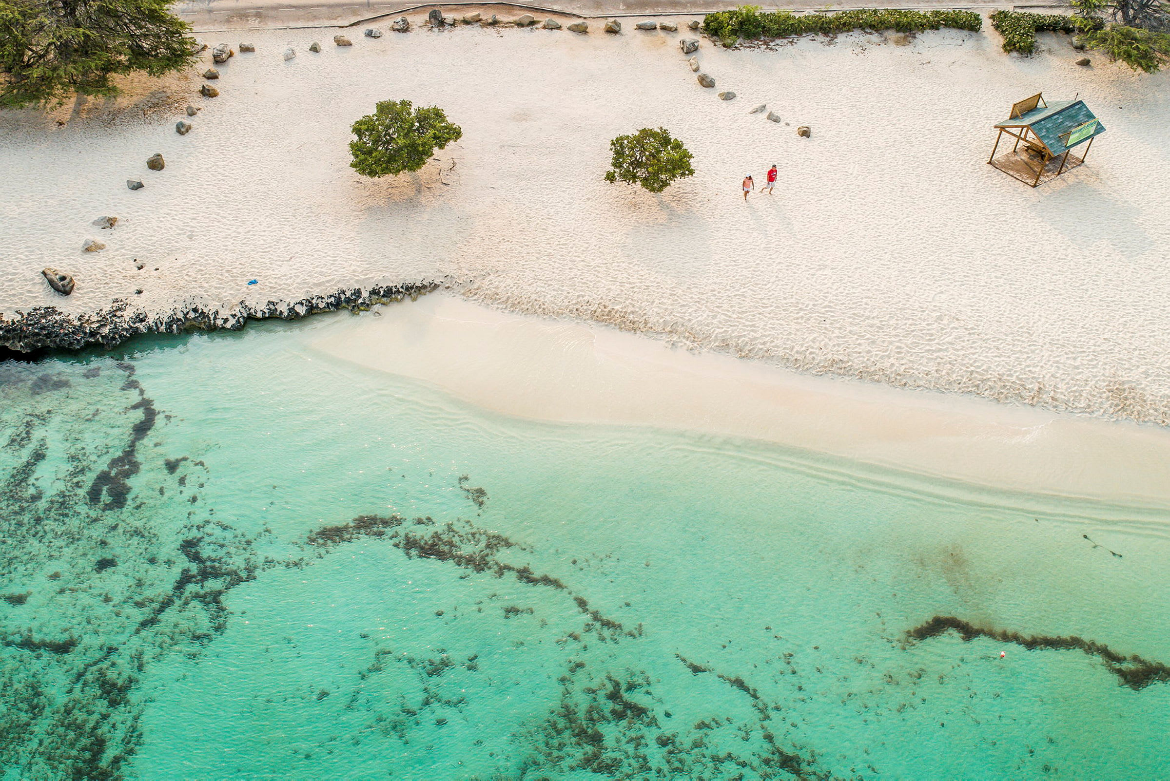 En strand med vit sand, gröna buskar och klart turkost vatten syns på bilden. Två människor går på stranden bredvid en liten trähytt. (AI-genererad text)