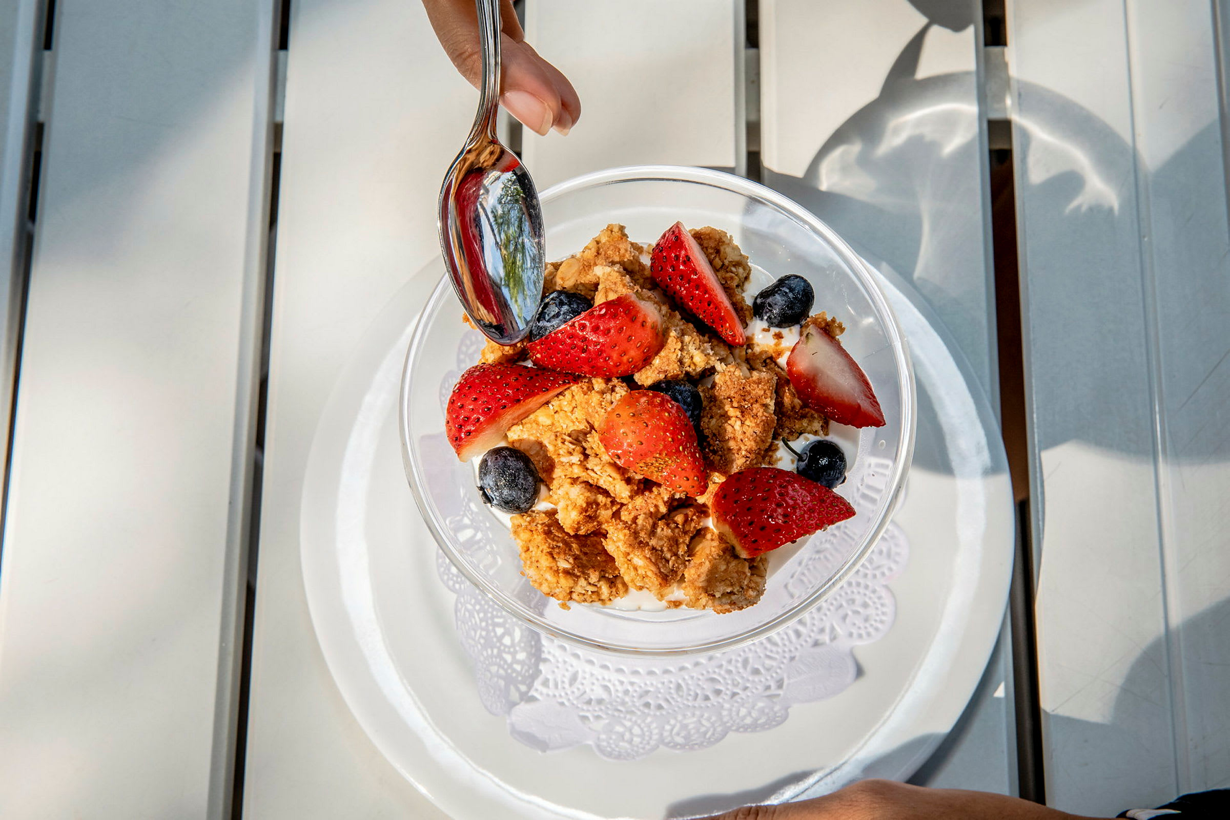 En skål med yoghurt, färska jordgubbar, blåbär och krispigt granola. En hand håller en sked ovanför maten. (AI-genererad text)