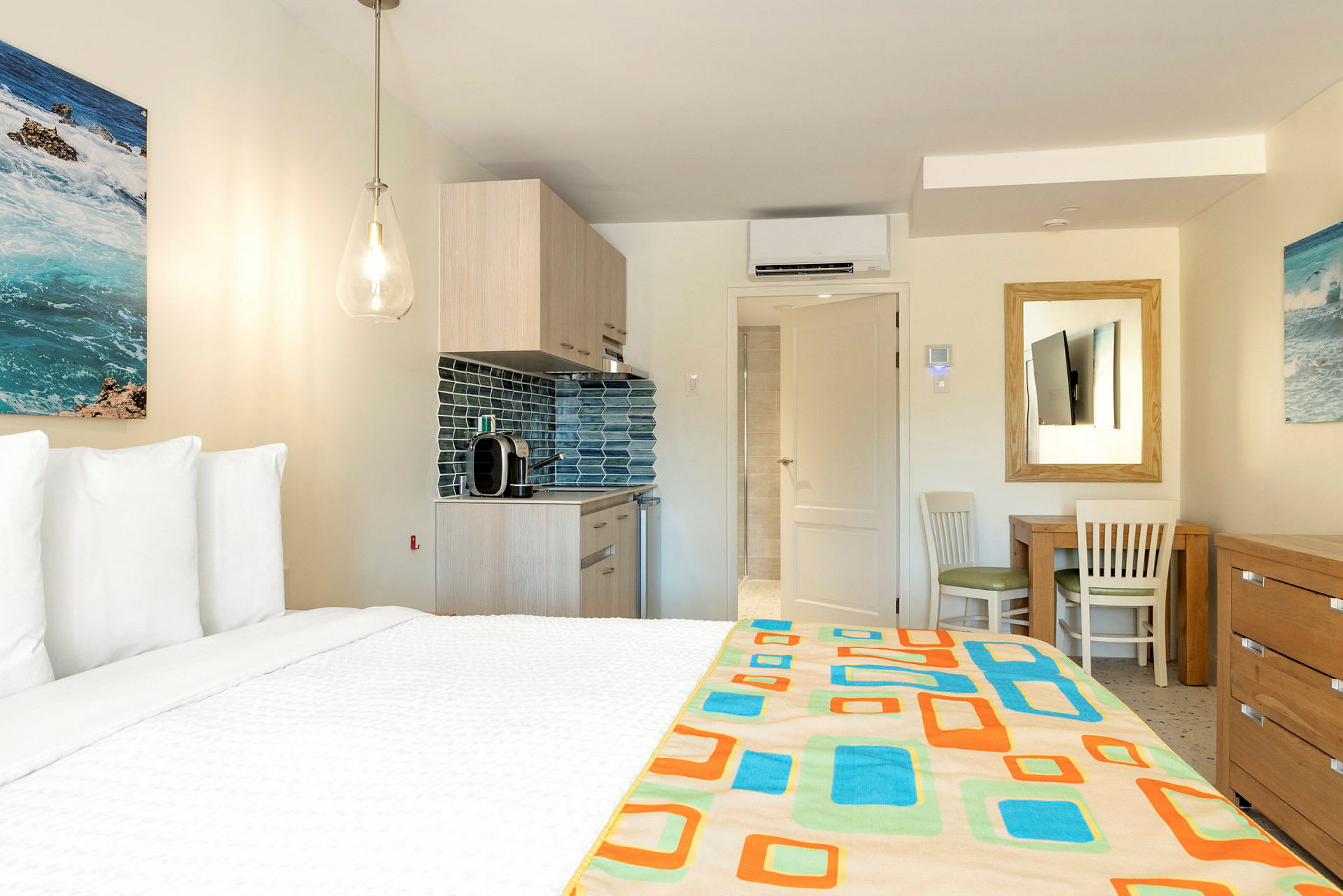 Ett hotellrum med en säng i förgrunden, ett kök och ett bord med stolar mot bakgrunden. Väggarna är dekorerade med målningar. (AI-genererad text)