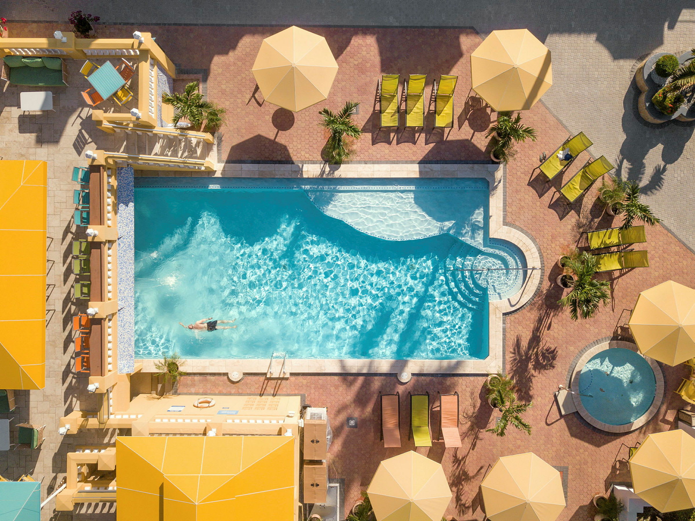 Uppifrån av en pool med en person som simmar. Runt poolen finns solstolar, parasoller och ett mindre bad. (AI-genererad text)
