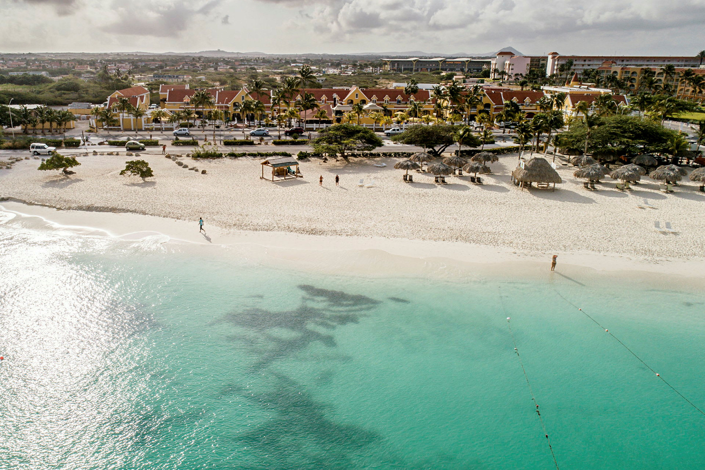 Sandstrand med turkost vatten, parasoller och några personer på stranden, samt byggnader i bakgrunden. (AI-genererad text)