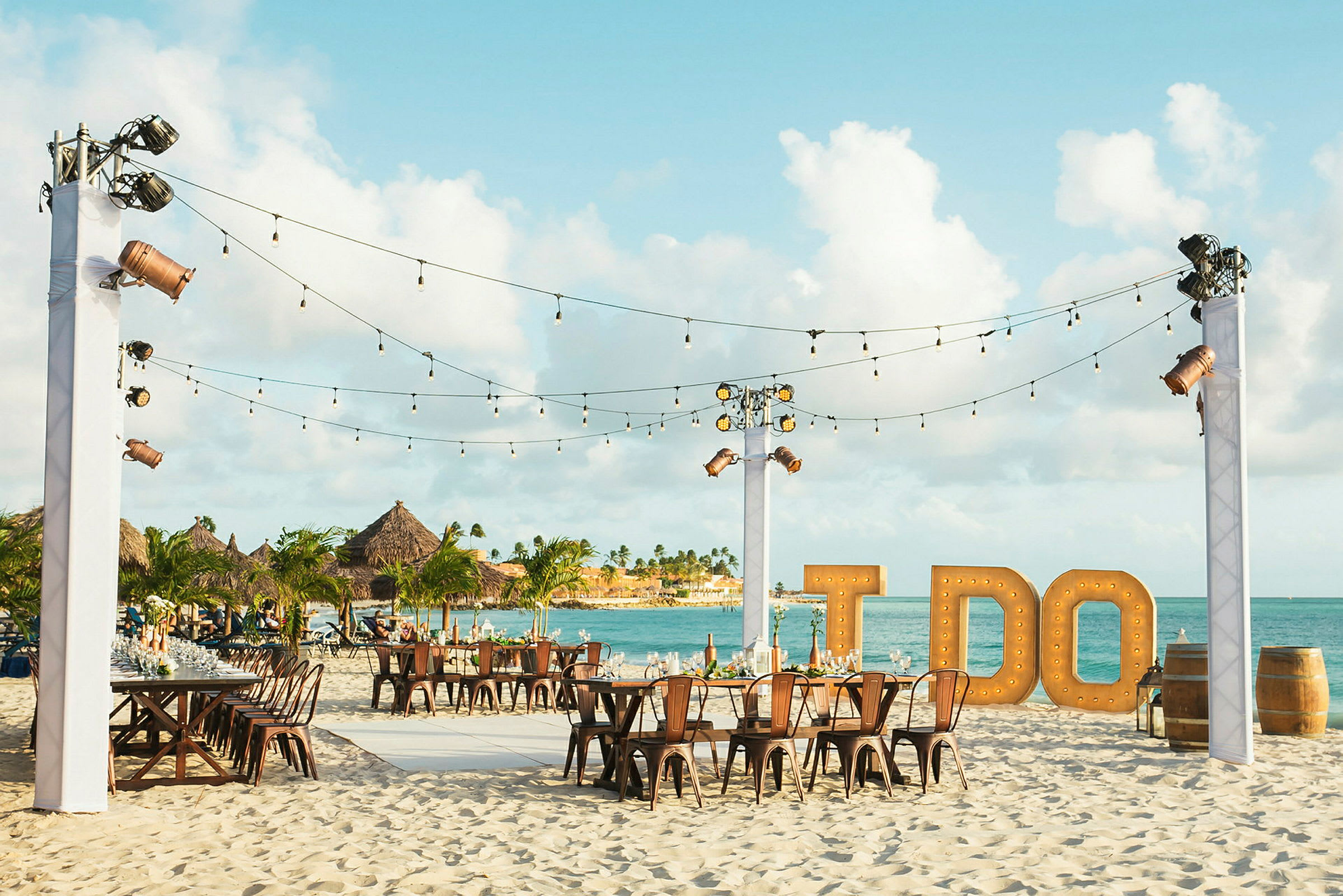 Ett strandbröllop med bord och stolar arrangerade på sanden, dekorationer med ljusslingor och stora bokstäver som bildar texten 'I DO' i bakgrunden. (AI-genererad text)