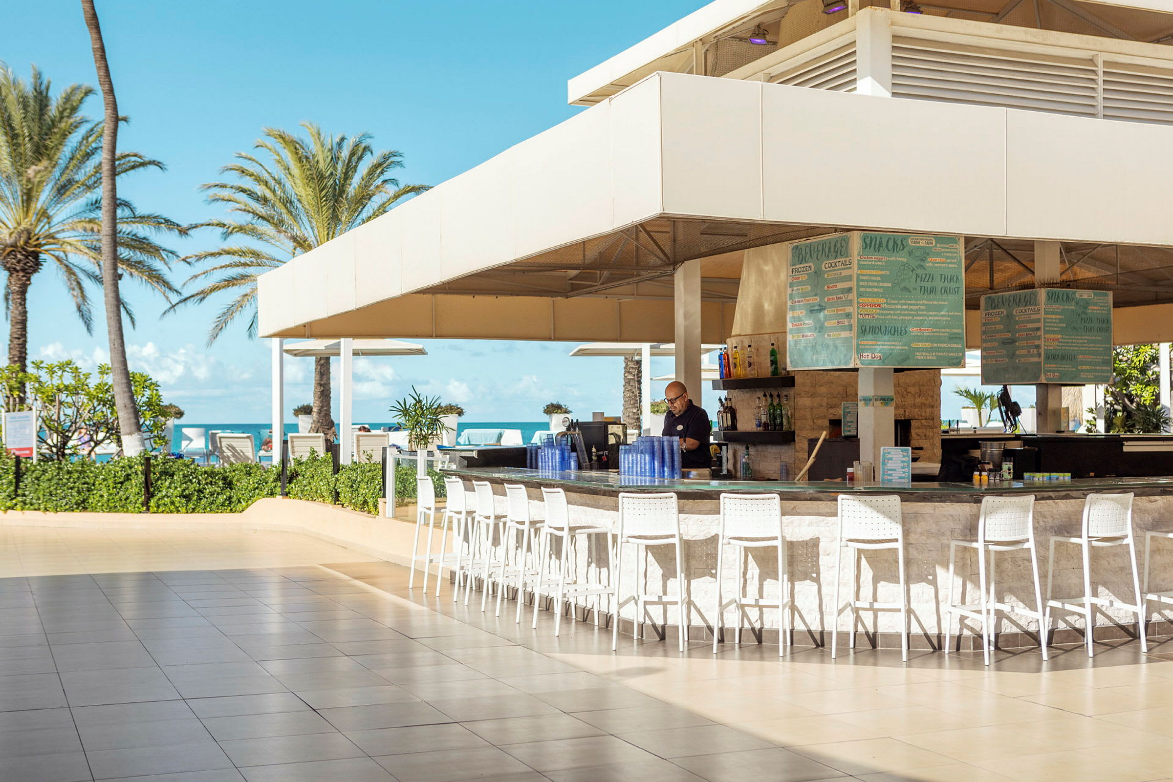 En bar utomhus med tak och höga stolar, belägen nära stranden med palmer och blå himmel i bakgrunden. (AI-genererad text)